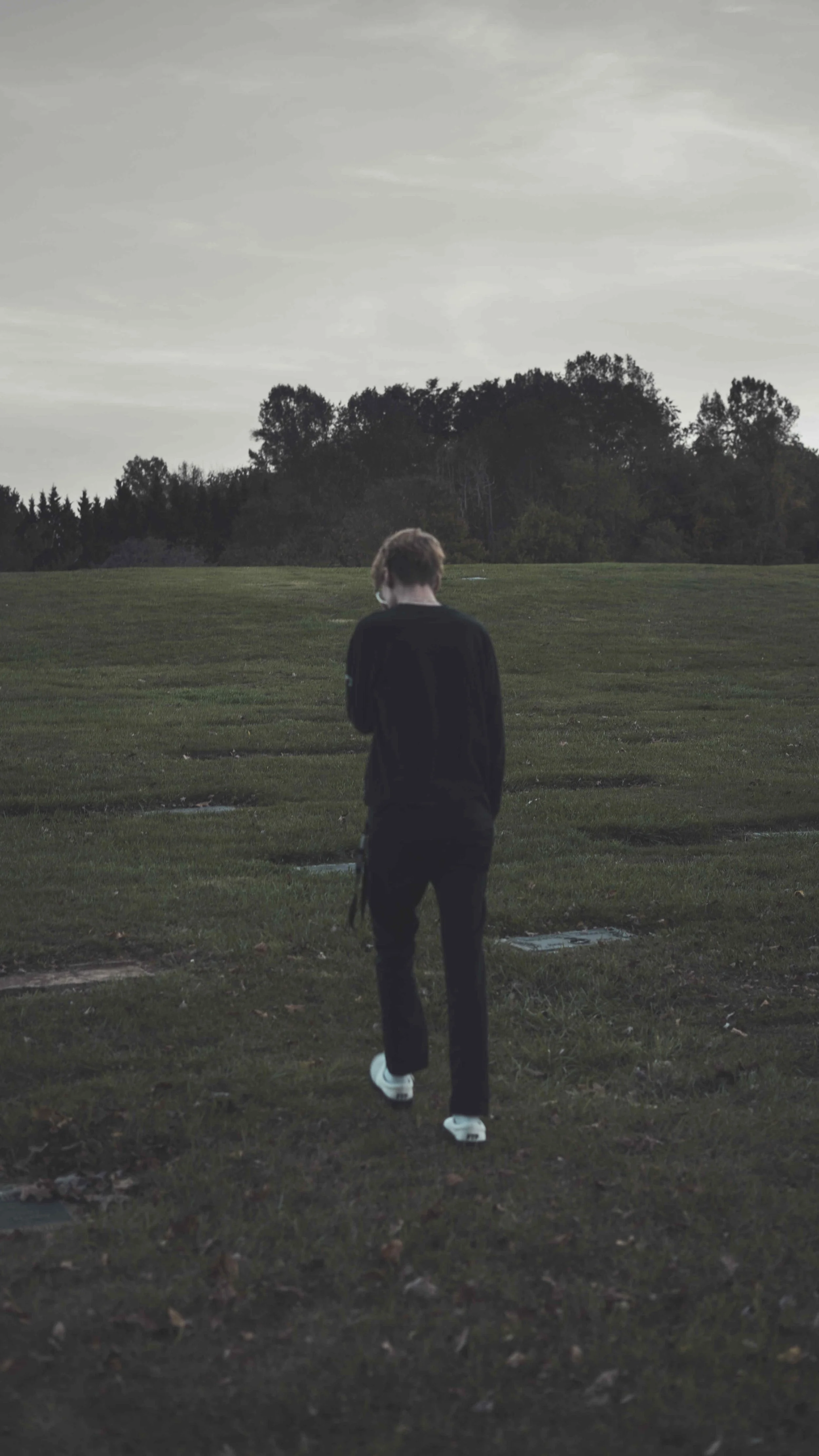A person with light brown hair walking on a grassy field toward a line of trees in the distance under an overcast sky.