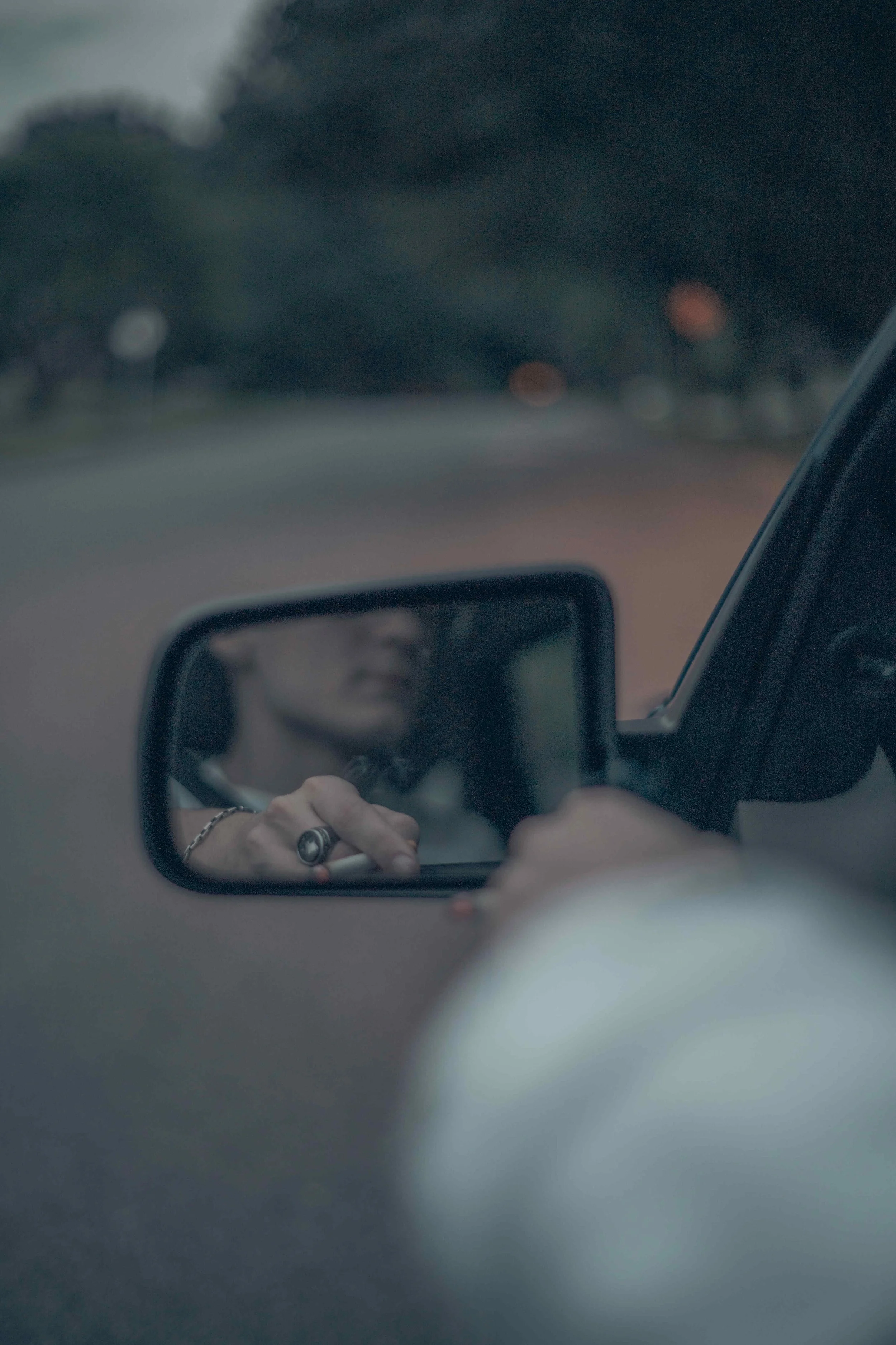 Close-up of a person's hand with rings holding a phone, reflected in a small rectangular mirror, with a blurred background of trees and sky seen through a vehicle window.