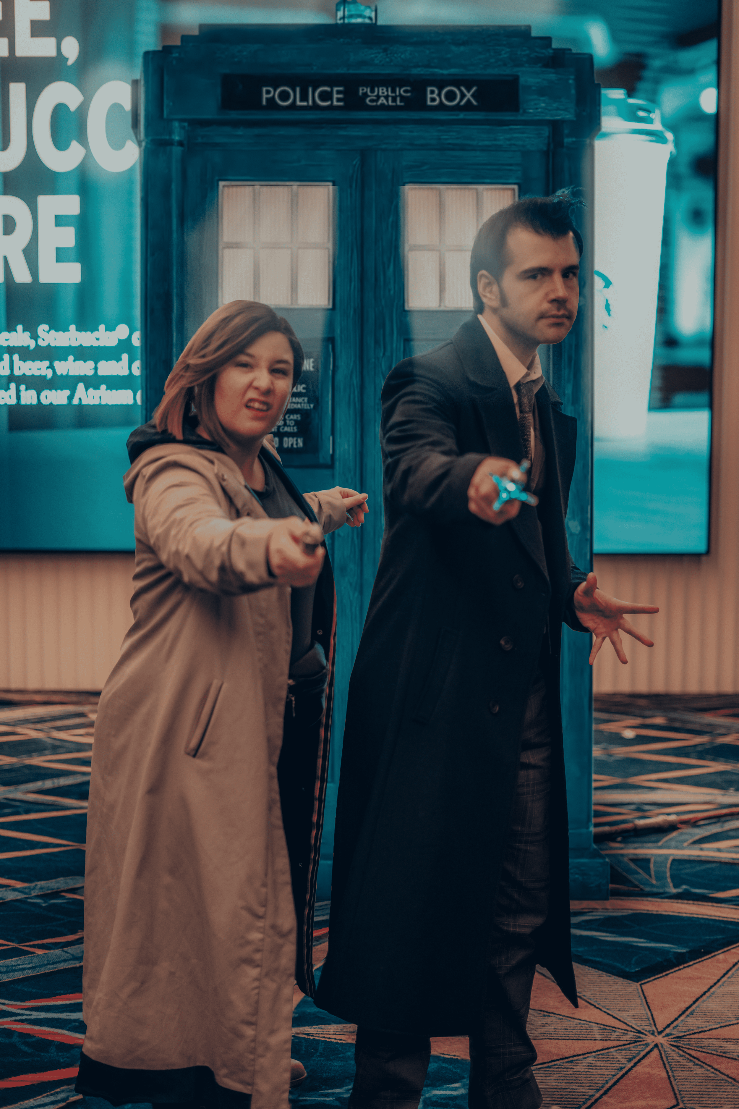 Two people pointing guns at the camera, standing in front of a blue police public call box and a white pillar. The woman on the left is wearing a beige trench coat, and the man on the right is dressed in a dark coat.