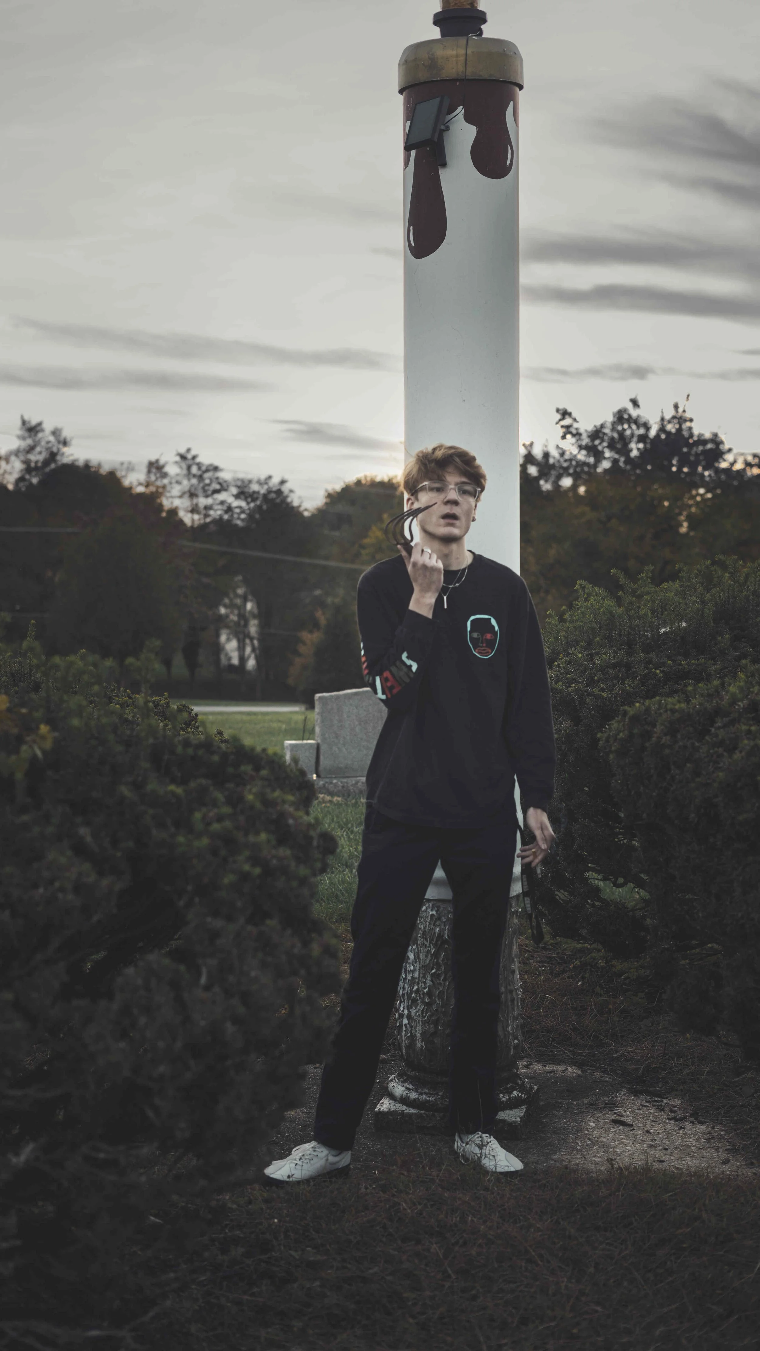 A young man with red hair, glasses, and casual clothing standing outdoors near a painted utility pole with a stormy sky in the background.