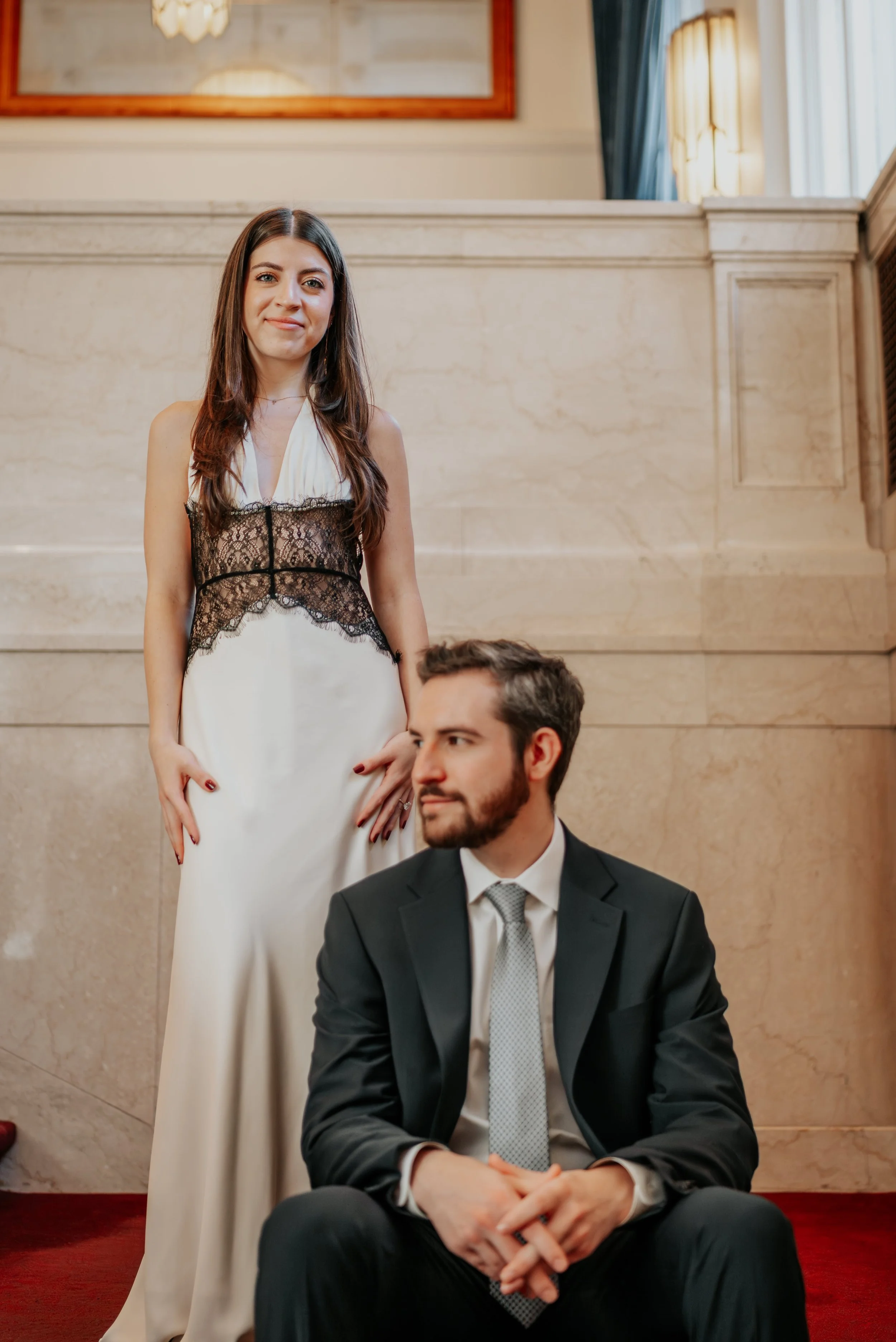 A woman in a white dress with black lace details standing behind a seated man in a black suit, white shirt, and gray tie, inside a formal building with marble walls and red carpet.