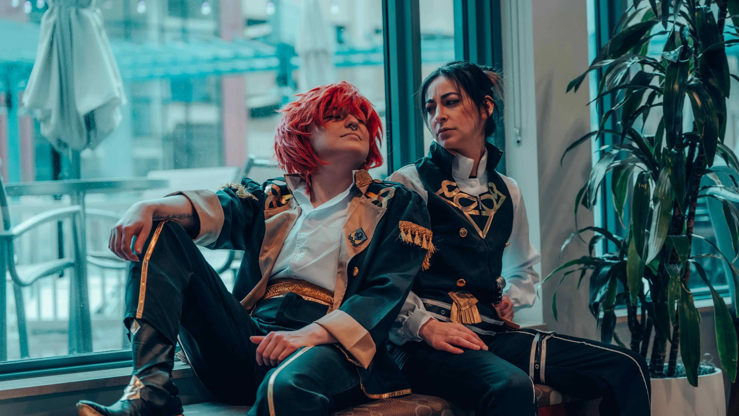 Two women sitting inside near a large window. One woman with short red hair and tattoos on her arm, dressed in a leather jacket with gold details, looks at the other woman with dark hair and a serious expression, who is also dressed in a black and wh