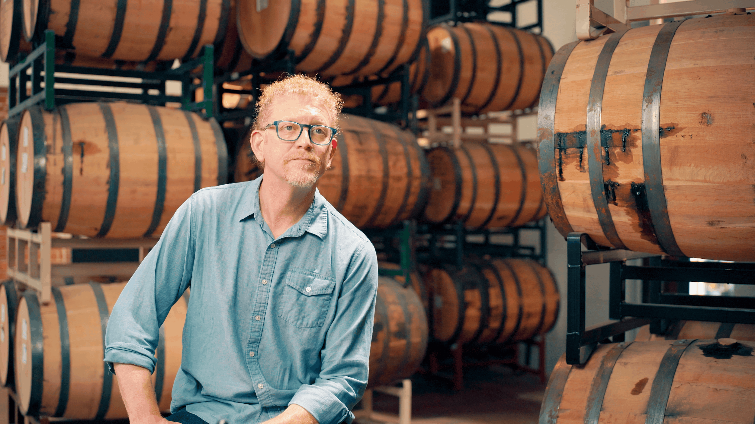 A man with glasses and a blue shirt standing in front of stacked wooden barrels in a cellar or storage room.