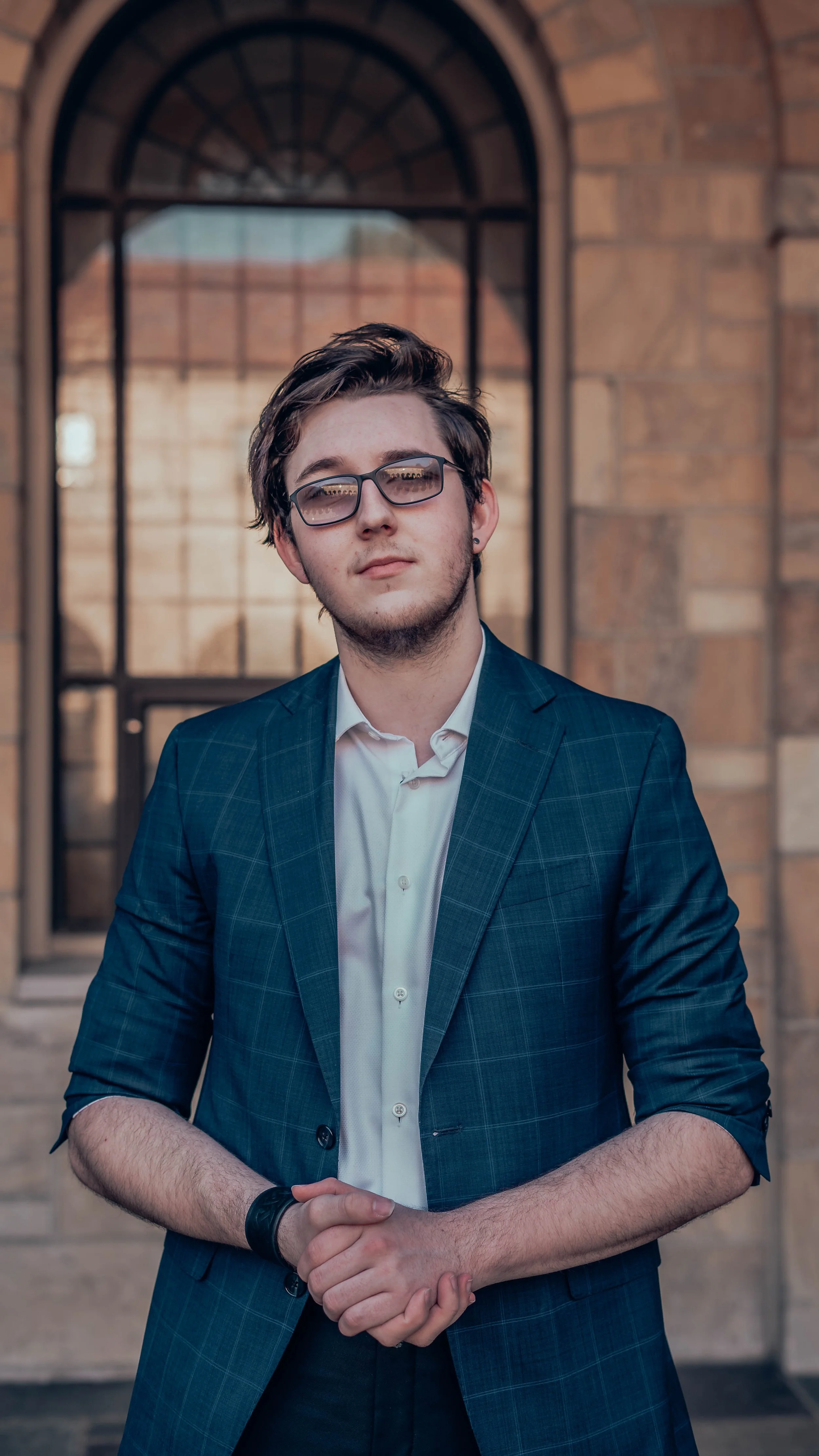 A young man dressed in a business suit and glasses standing outdoors in front of a brick building with a large arched window.