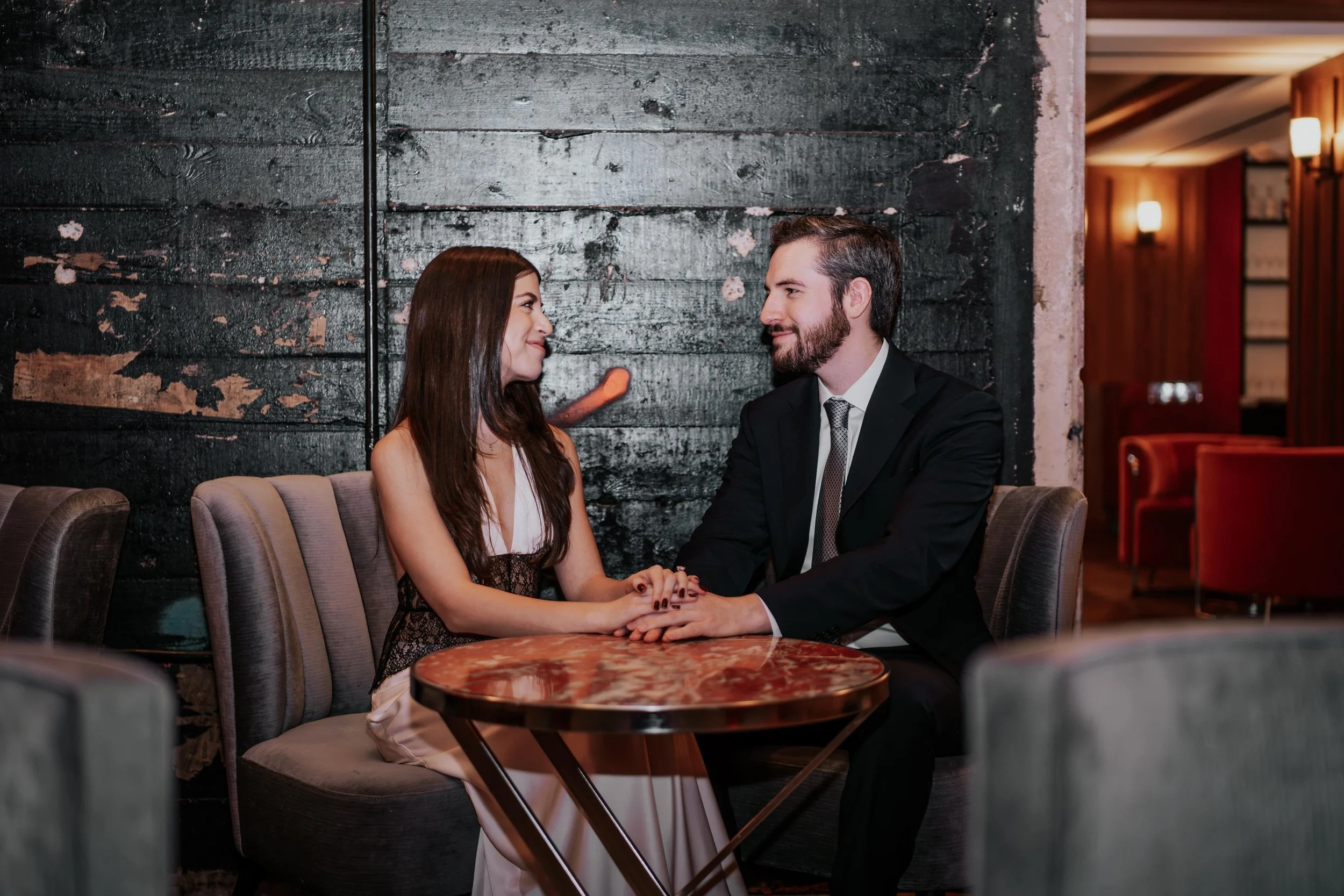 A man and woman sitting at a table, holding hands and gazing at each other in a dimly lit restaurant.
