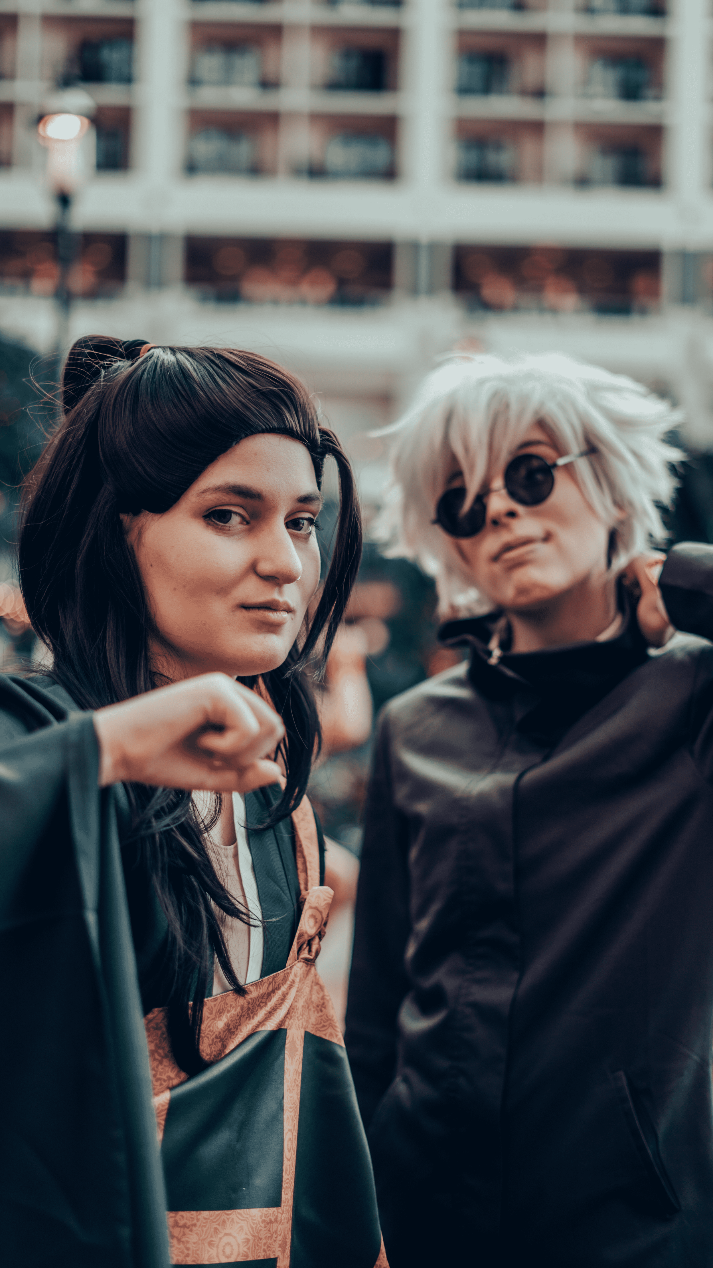 Two people in costumes, one with dark hair and one with white hair and round sunglasses, standing outdoors with a building in the background.