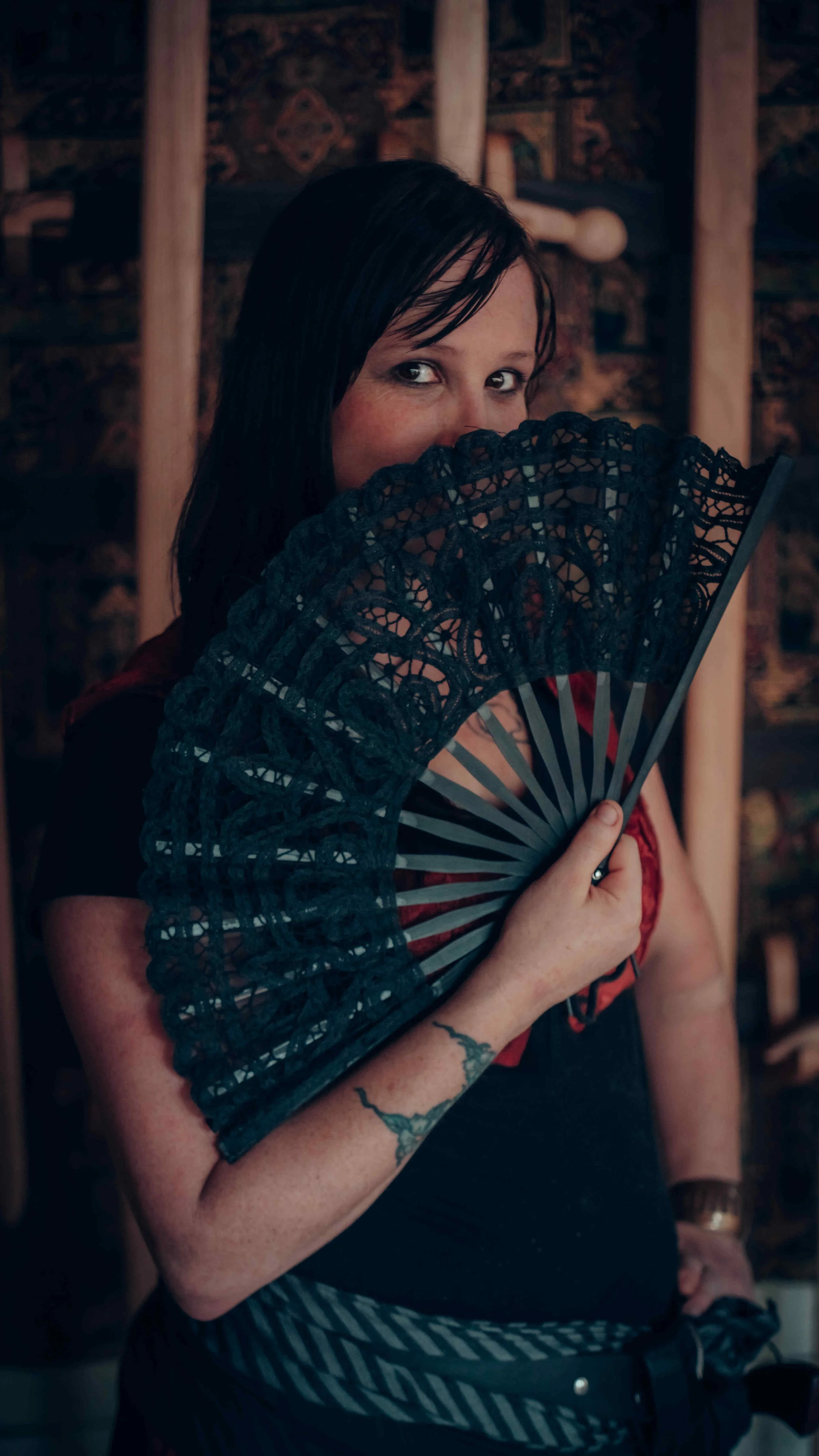 A woman with dark hair holding a black lace fan partly covering her face, looking at the camera with a sly expression, in a dimly lit room with wooden beams and patterned wallpaper.