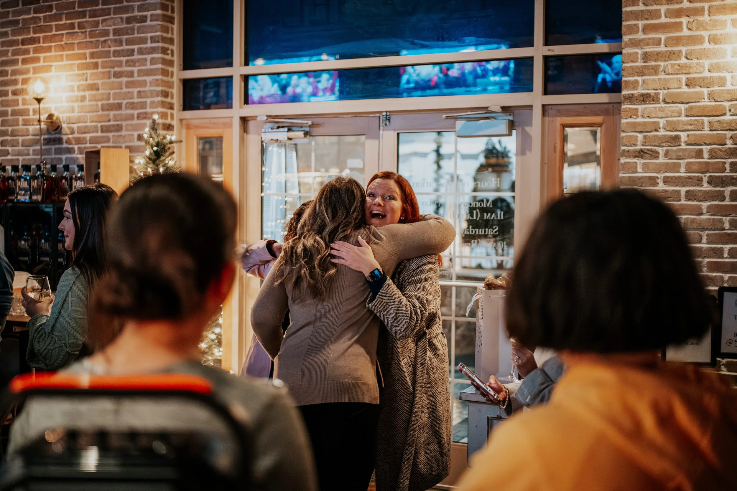 Two women are hugging each other inside a restaurant, with a woman in a beige coat smiling happily. Other diners are seated around them, and a Christmas tree with lights is visible in the background. The interior has brick walls and large windows.