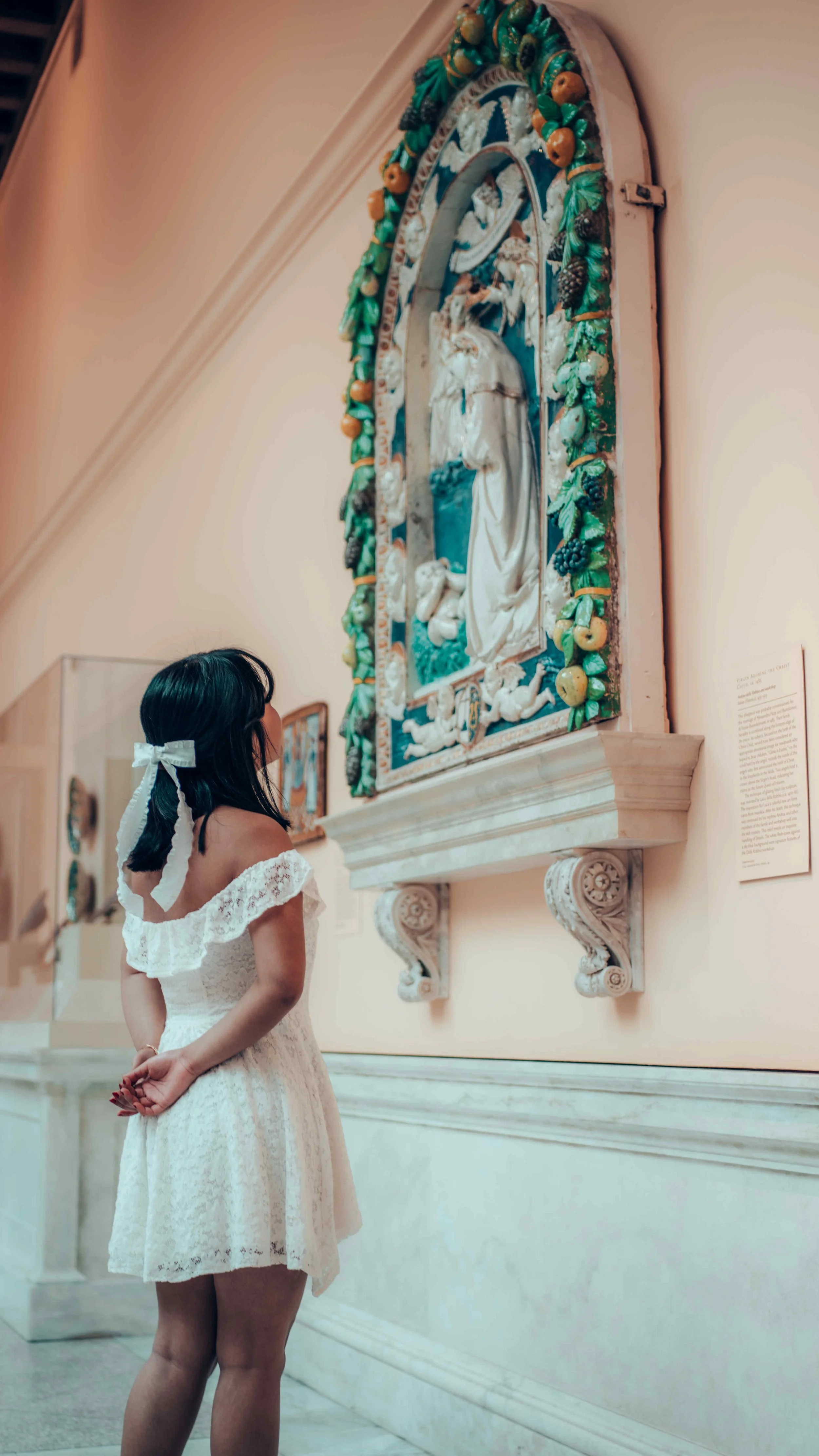 A young girl with a white lace dress and a ribbon in her hair looking at a religious painting in a museum.