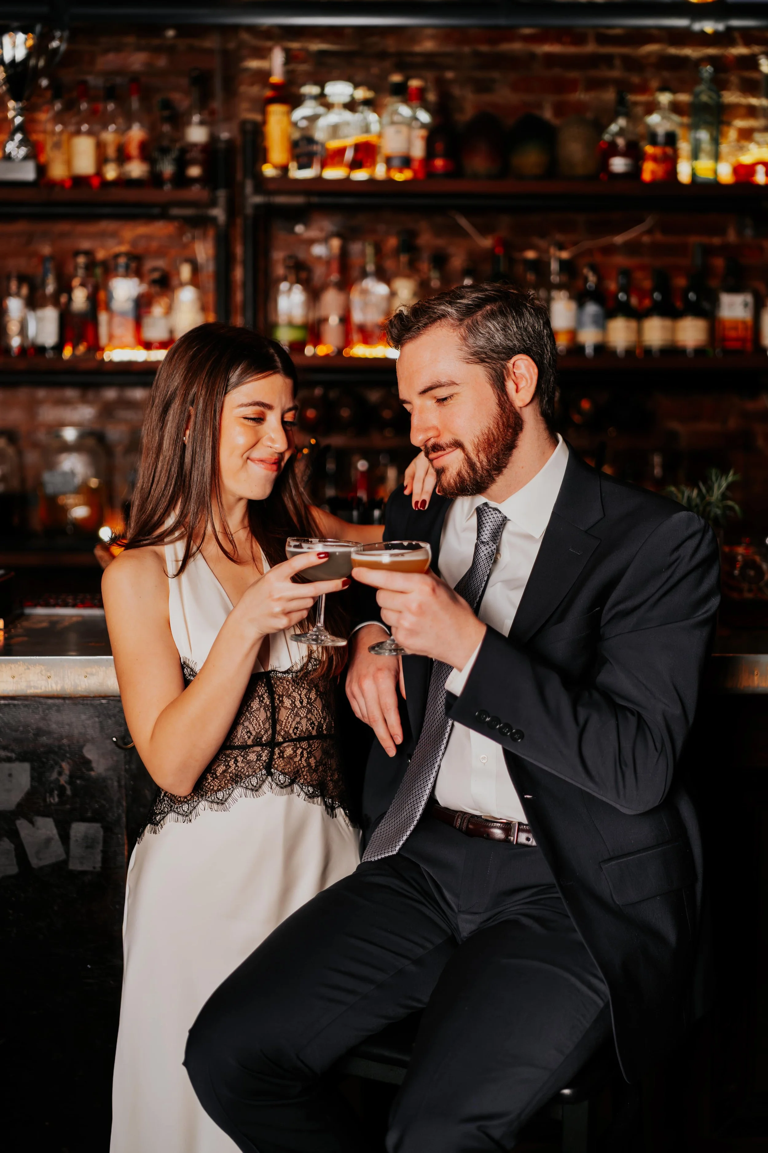 A man and woman in formal attire sharing a toast with cocktails at a bar, smiling at each other, with a background of liquor bottles on shelves.