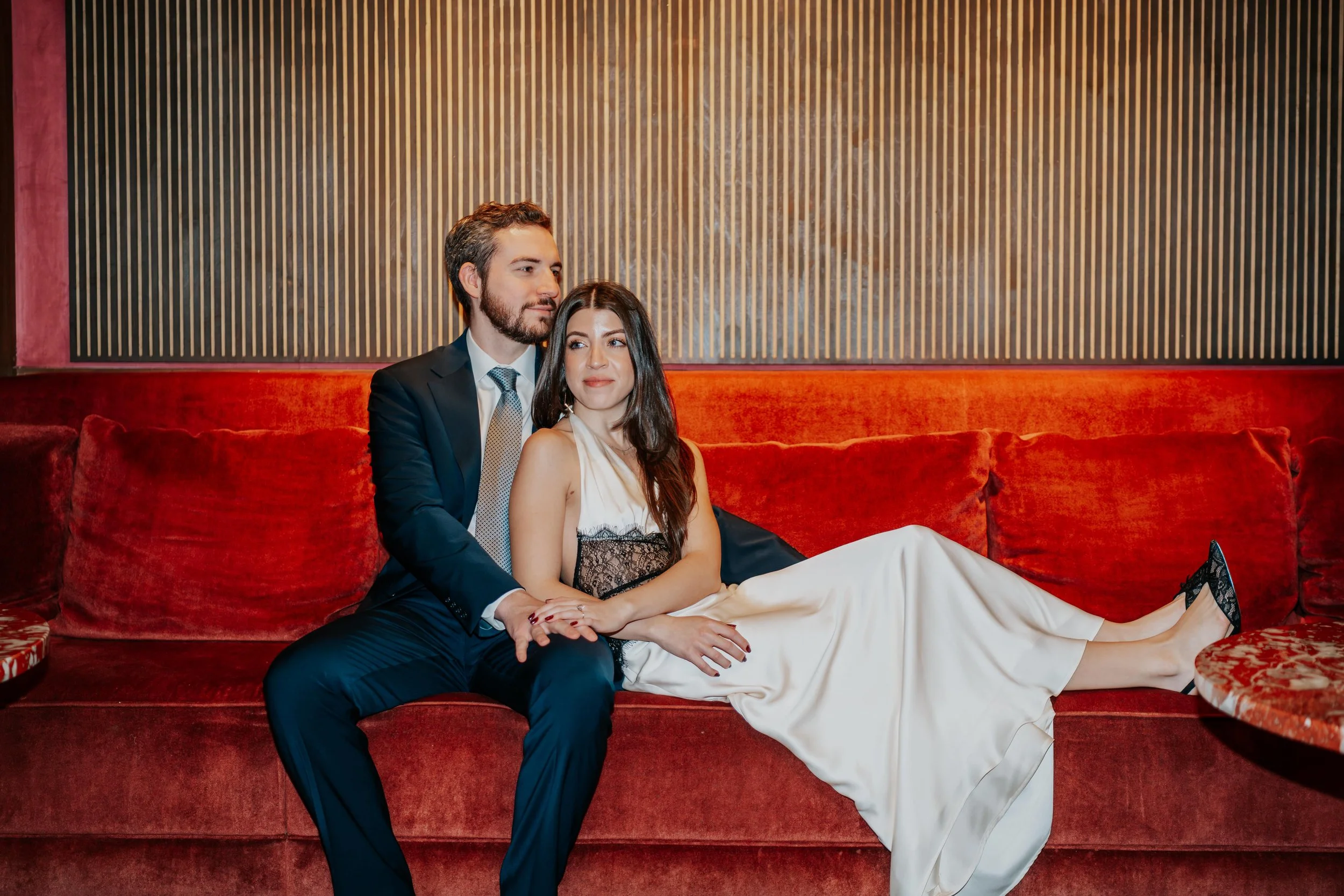 A man and a woman sitting on a red velvet couch in a cozy, warmly lit room. The man is wearing a navy suit with a white shirt and a patterned tie, and has his arm around the woman. The woman is in a white dress with black lace details, and she is wea