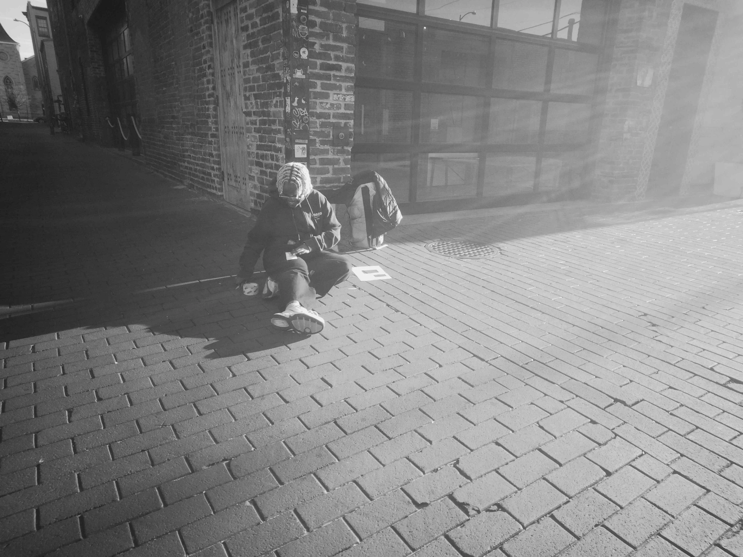 A person with blond dreadlocks sitting on a sidewalk outside a brick building, reading a pamphlet or card, with a backpack and a water bottle nearby, in daylight with shadows on the ground.
