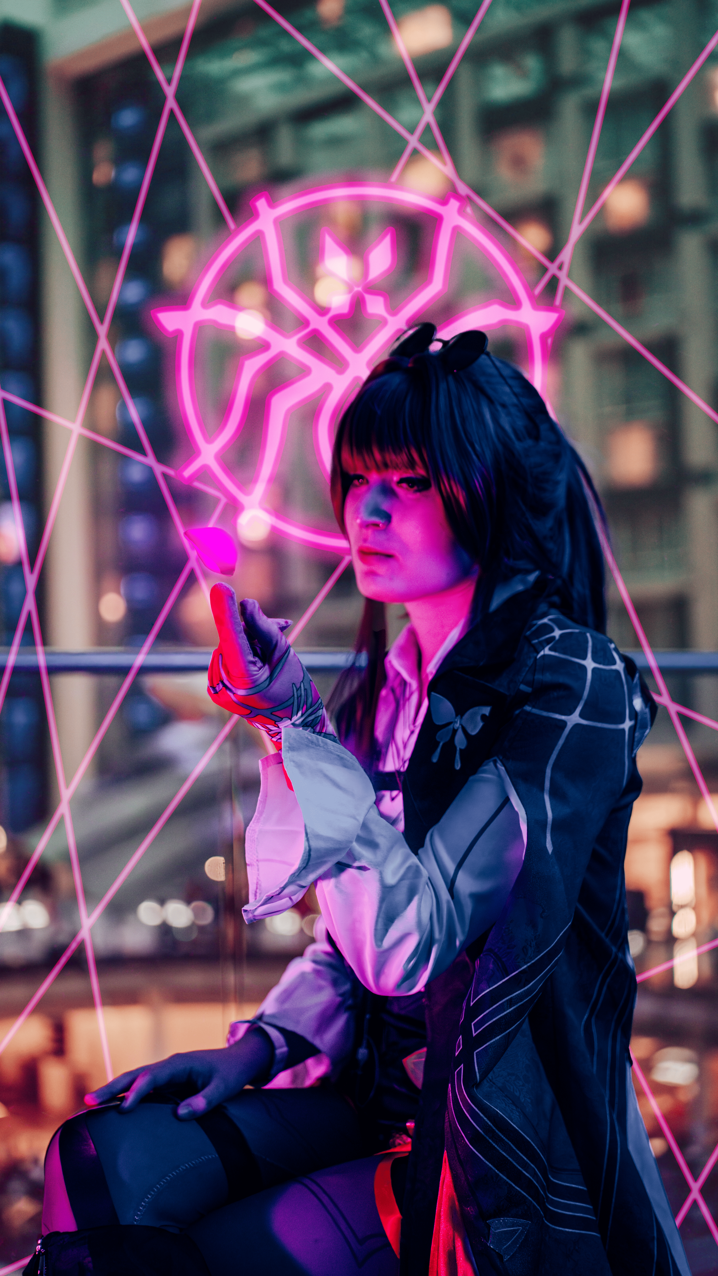 A woman with long black hair and sunglasses perched on her head posing in front of a pink neon butterfly symbol. She is wearing a black and white jacket and purple gloves. The background shows a building with illuminated windows, and the scene is lit