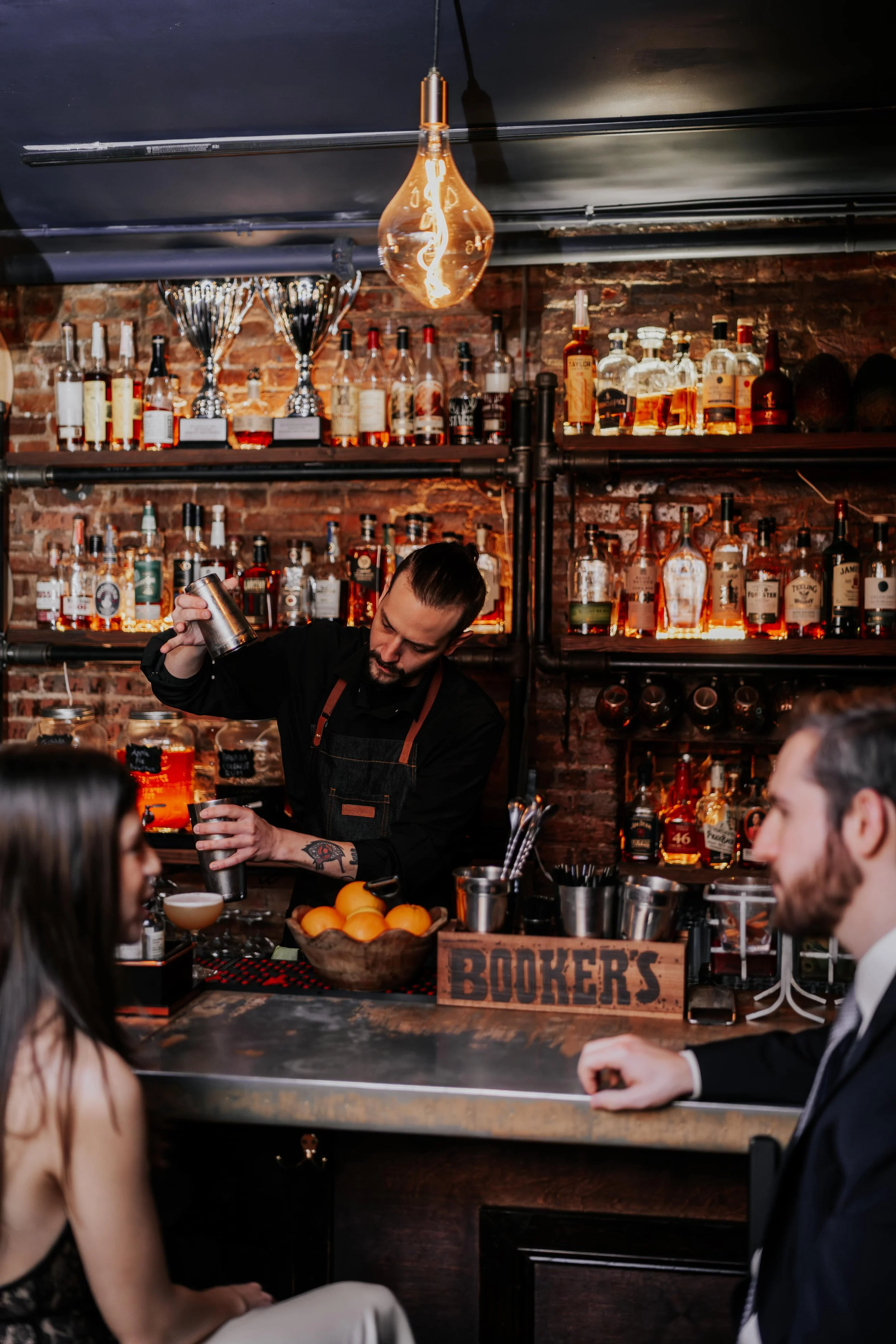 A bartender preparing a cocktail at a bar with liquor bottles on shelves behind him, while two customers, a woman and a man, sit at the bar watching him.