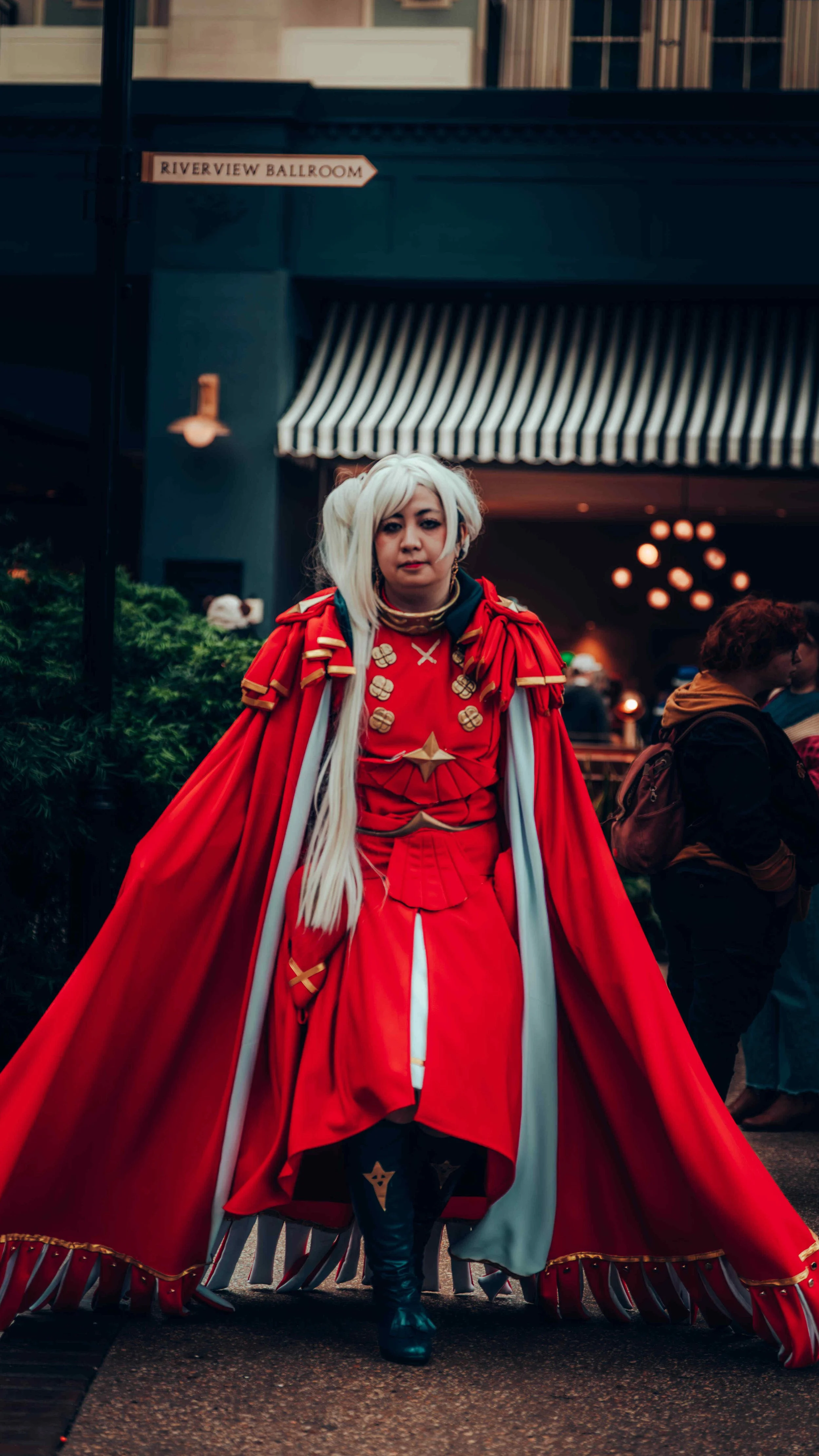 Person dressed in an elaborate red and white costume with long white hair, walking outdoors near a building with a striped awning and a sign that reads 'Riverview Ballroom'.