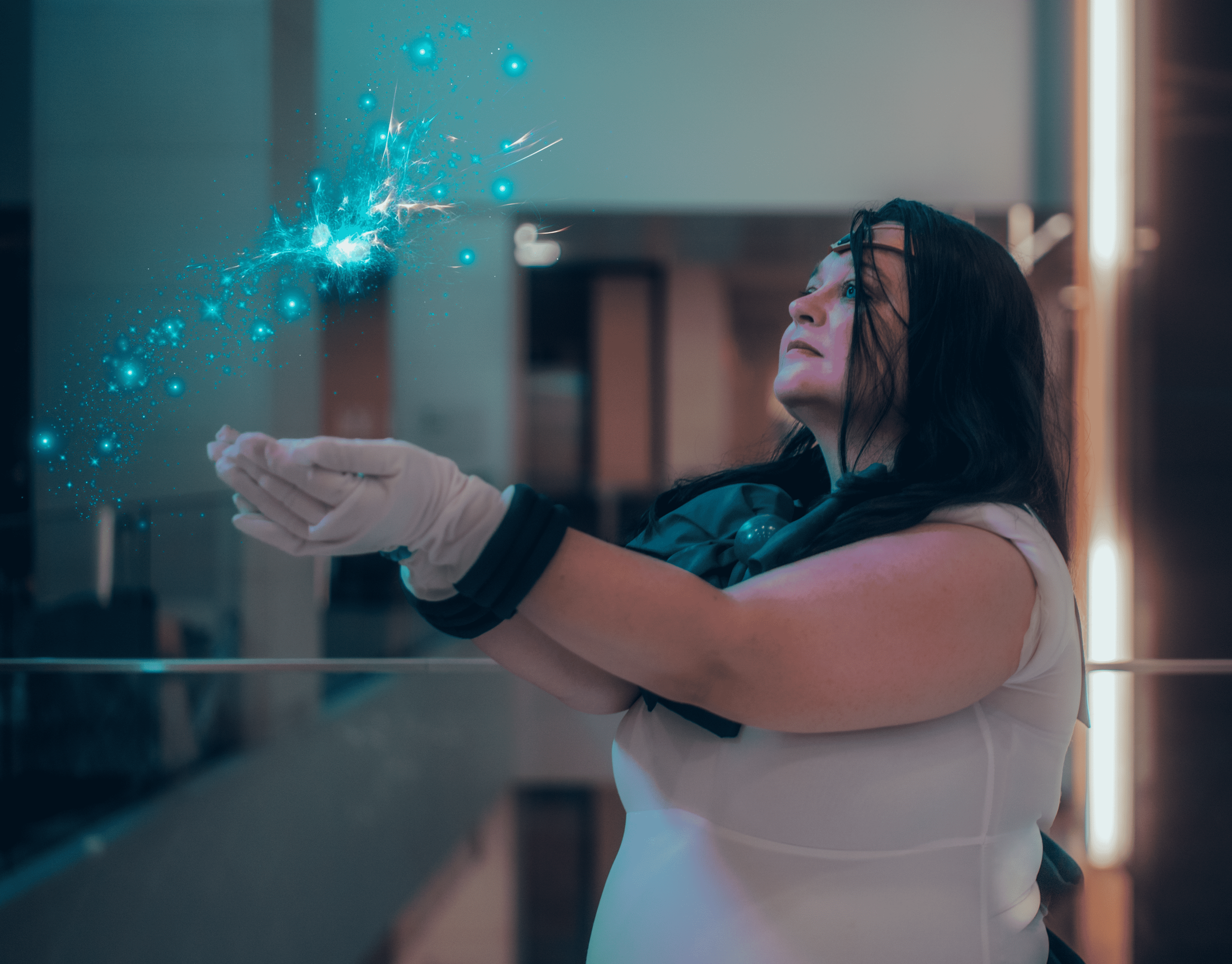 A woman with dark hair and glasses, wearing a white shirt, is in a room with modern decor. She is holding her hands apart as glowing blue digital particles and sparks are emanating from her palms, creating a futuristic visual effect.