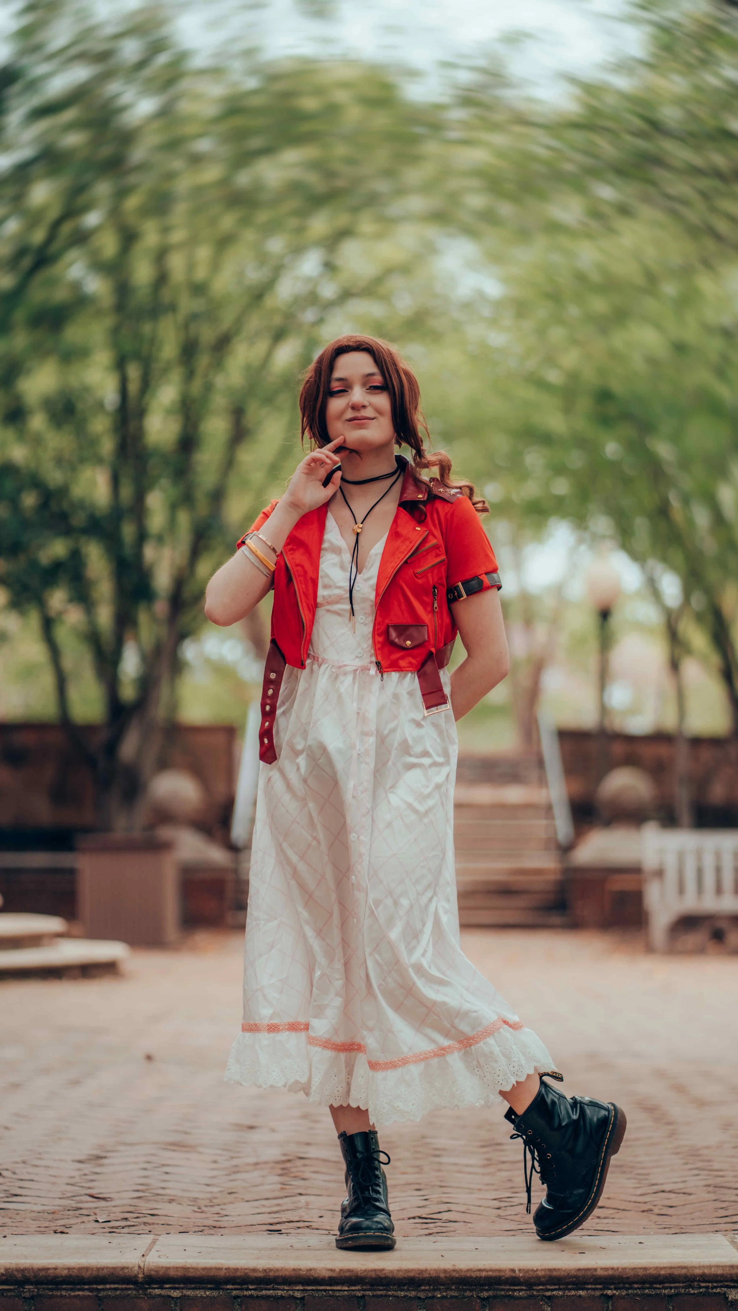 A young woman with brown hair and fair skin stands outdoors on a brick pathway, wearing a red jacket over a white dress with a pattern, black combat boots, and accessorized with necklaces and bracelets, with a background of trees and steps.
