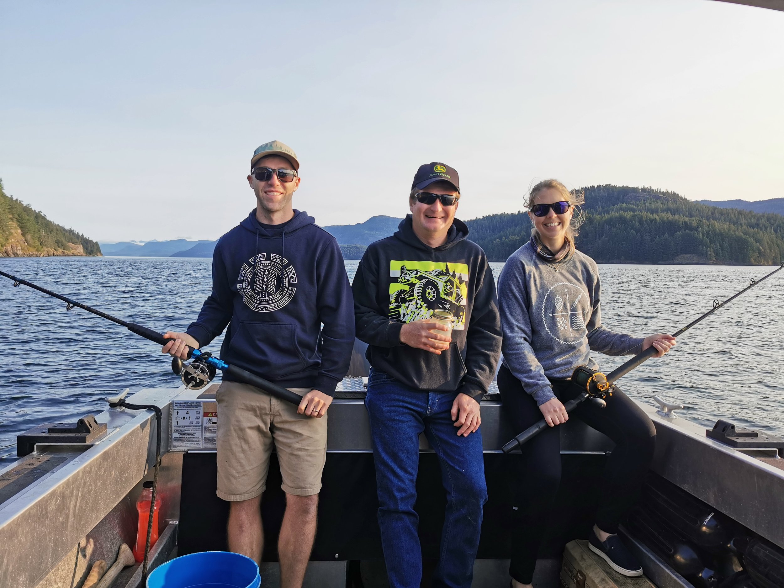 Three people sitting on a boat, holding fishing rods, with a scenic view of water and forested hills in the background.