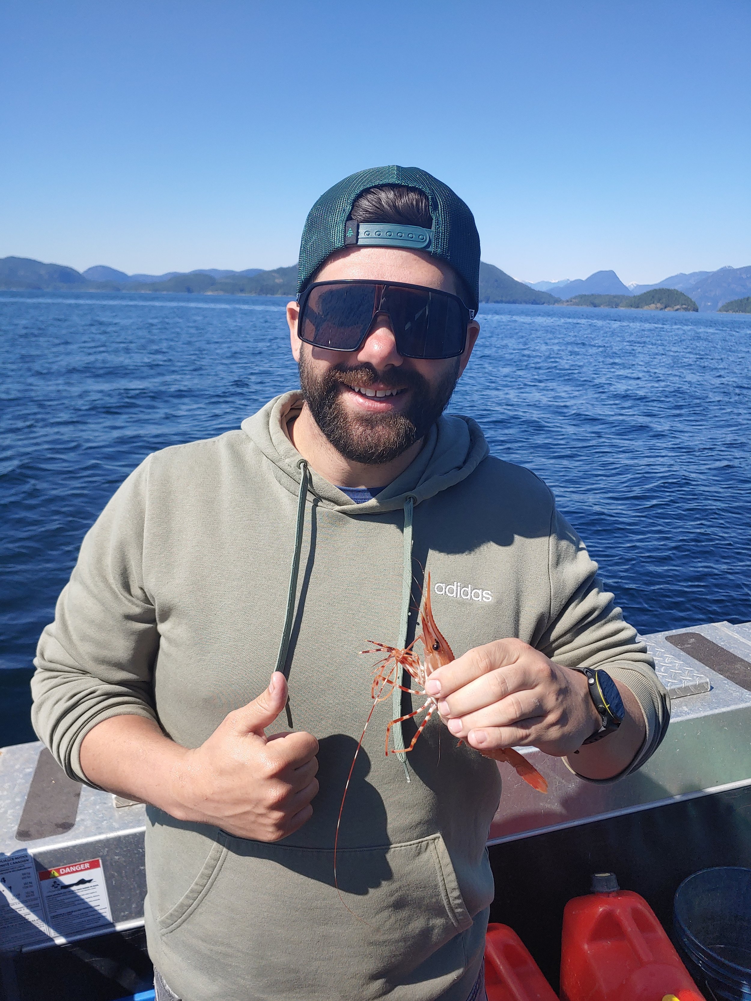 Person in sunglasses and cap holding a shrimp on a boat with ocean and mountains in the background.