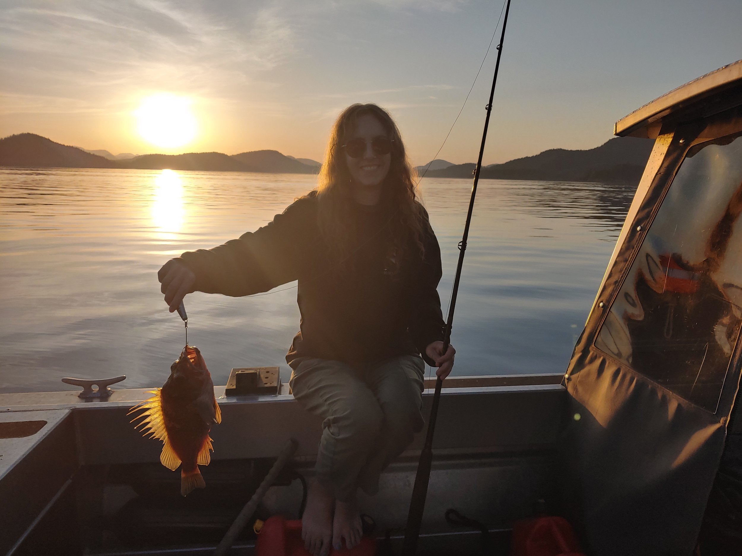 Person holding a fish on a boat during sunset with mountains and water in the background.