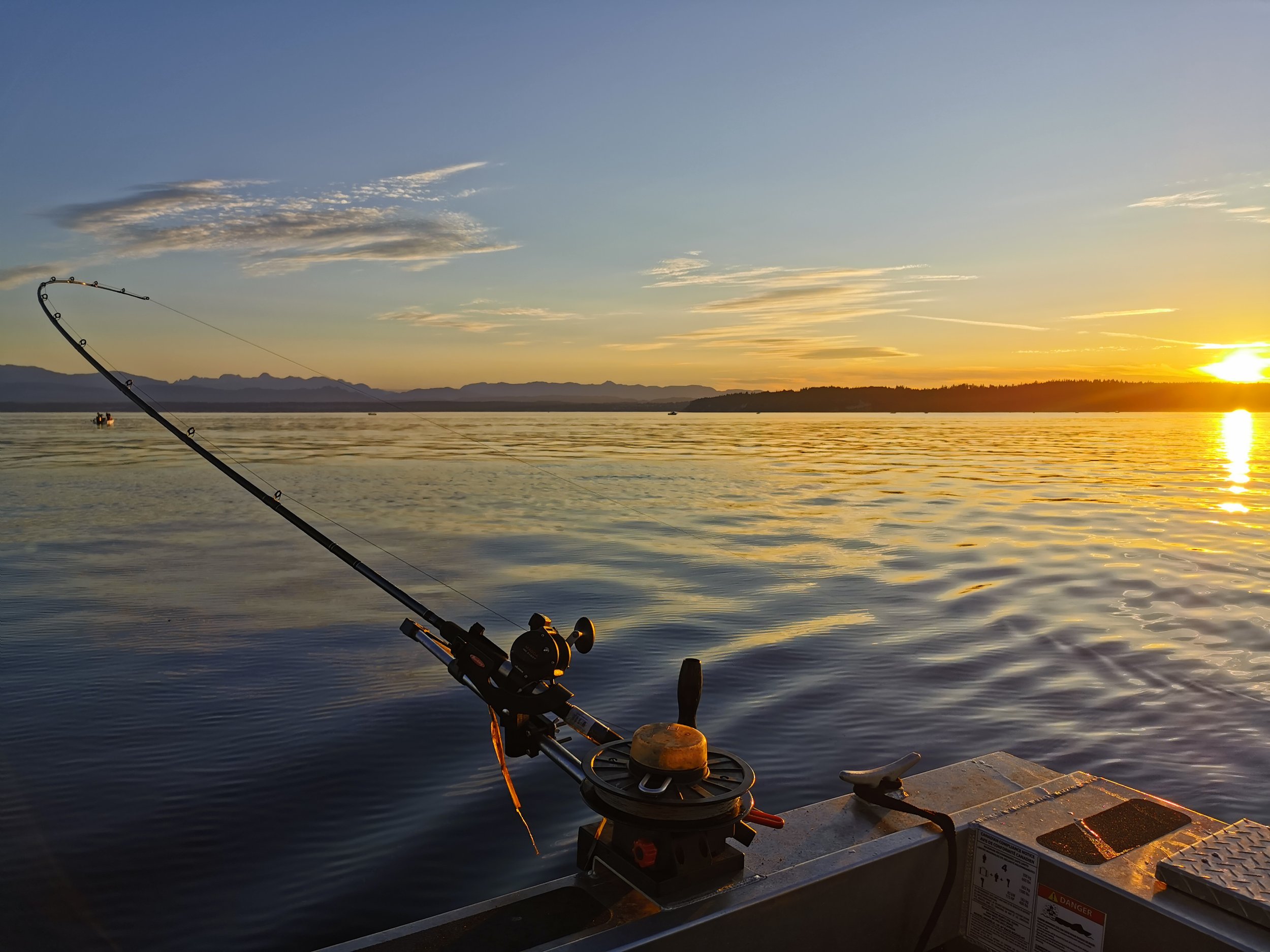 Fishing rod on a boat at sunset over calm ocean water with distant mountains and clear sky.