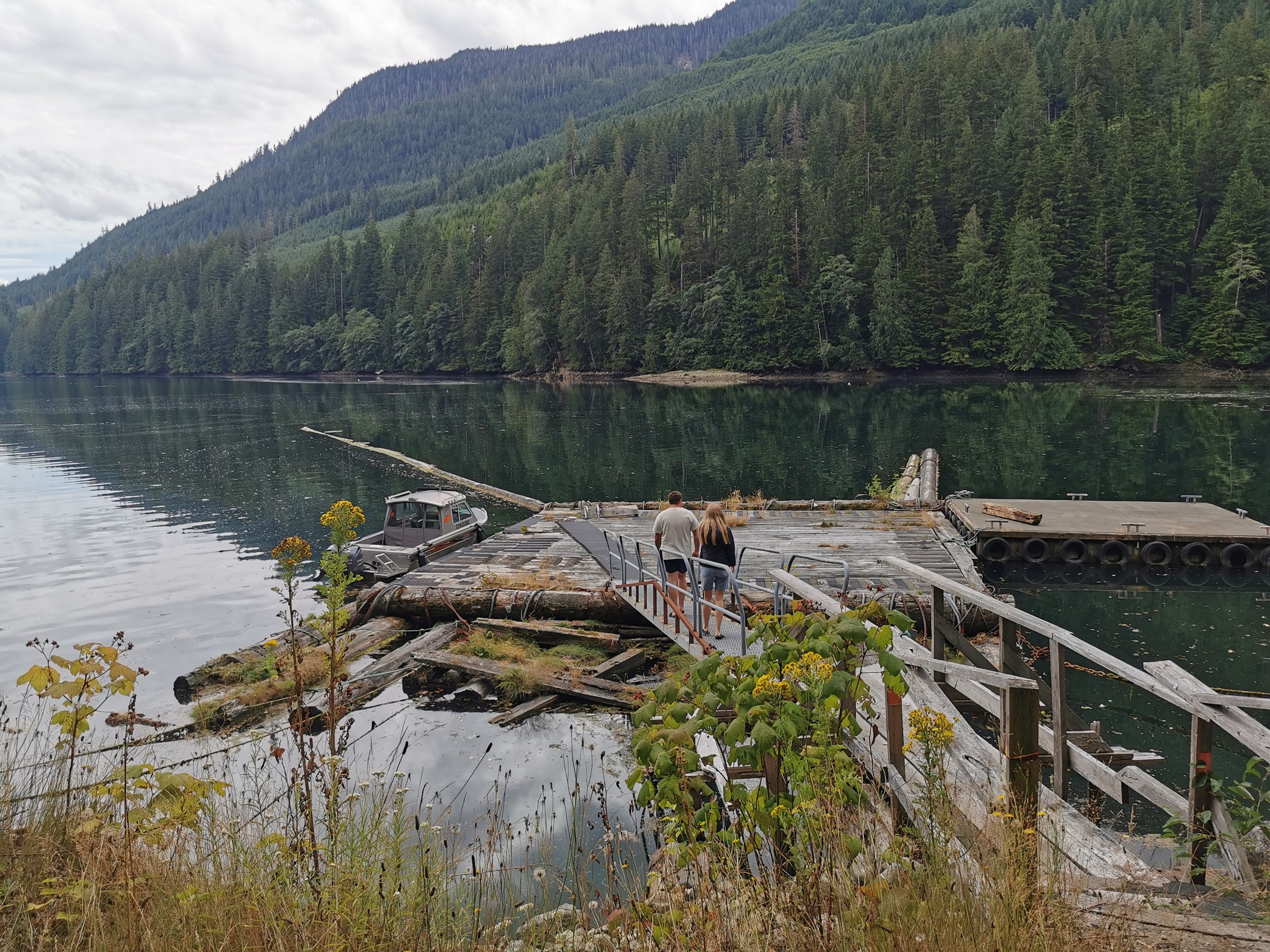Couple walking on a wooden dock by the pacific ocean with forested mountains in the background.