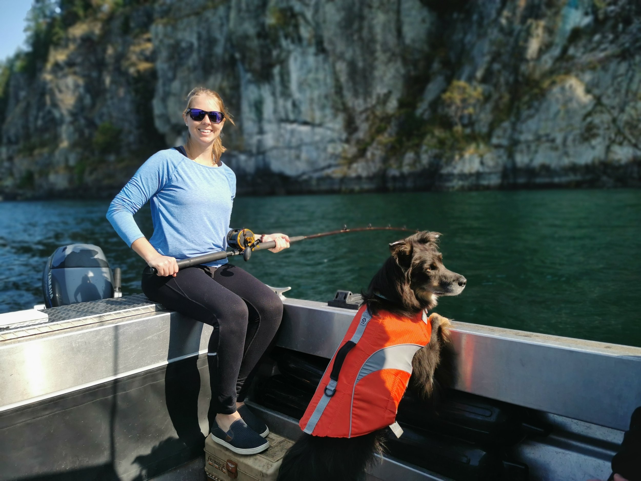 Woman sitting on a boat holding a fishing rod with a dog wearing an orange life jacket, scenic cliffs in the background.