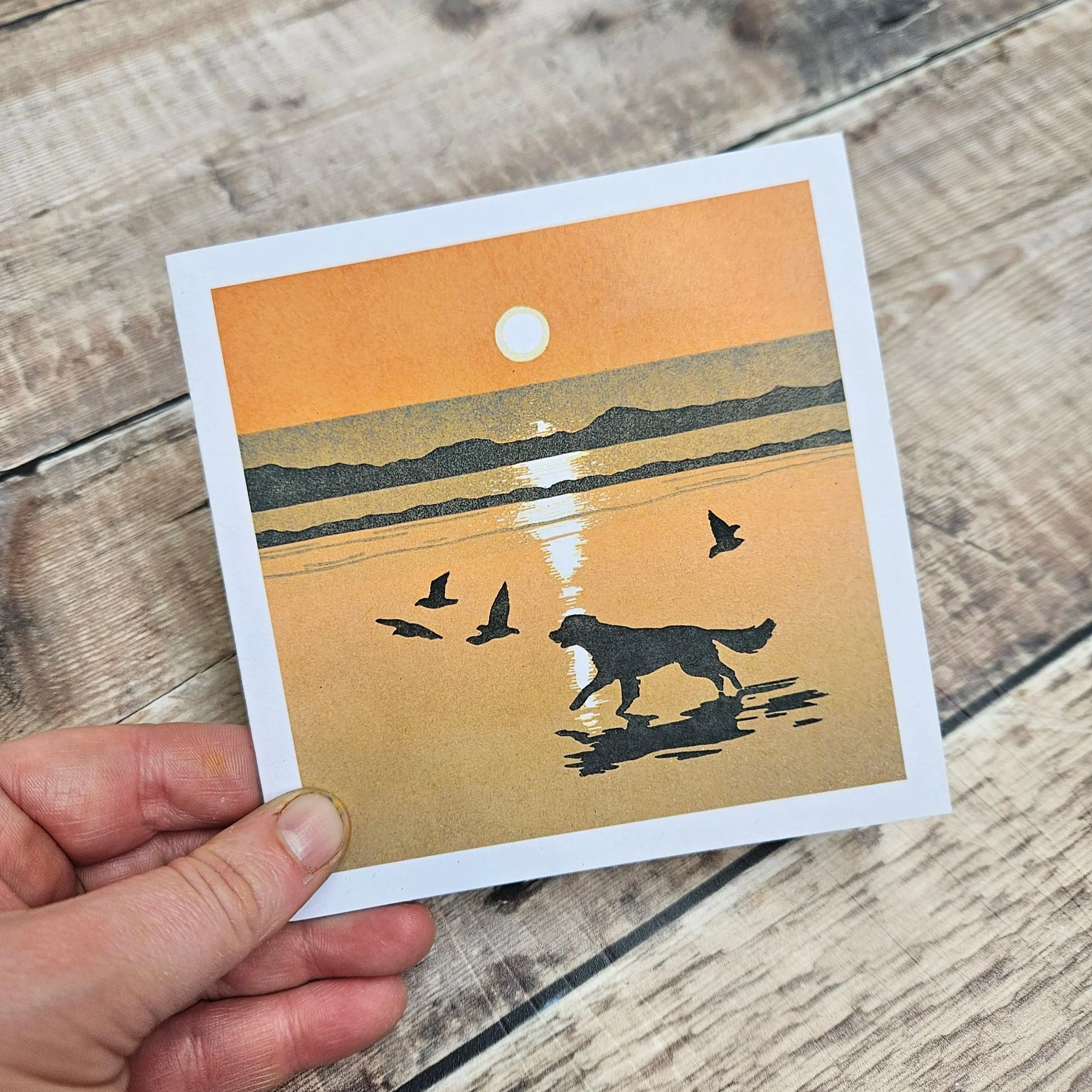 A hand holding a small print of a sunset scene with a dog chasing birds on a  beach.