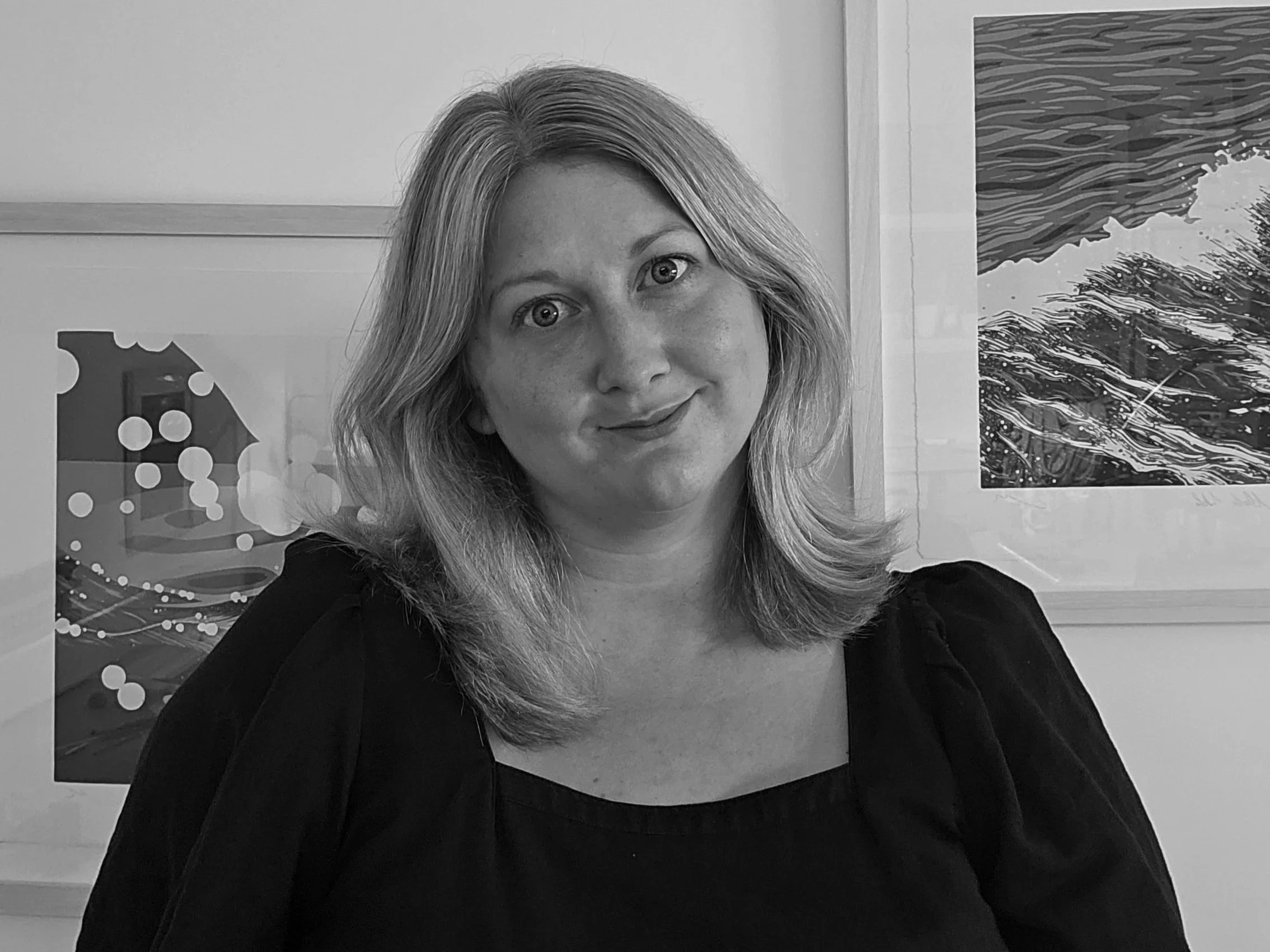 The artist Megan Hall with shoulder-length hair smiling, wearing a dark top, standing in front of framed artwork featuring waves.