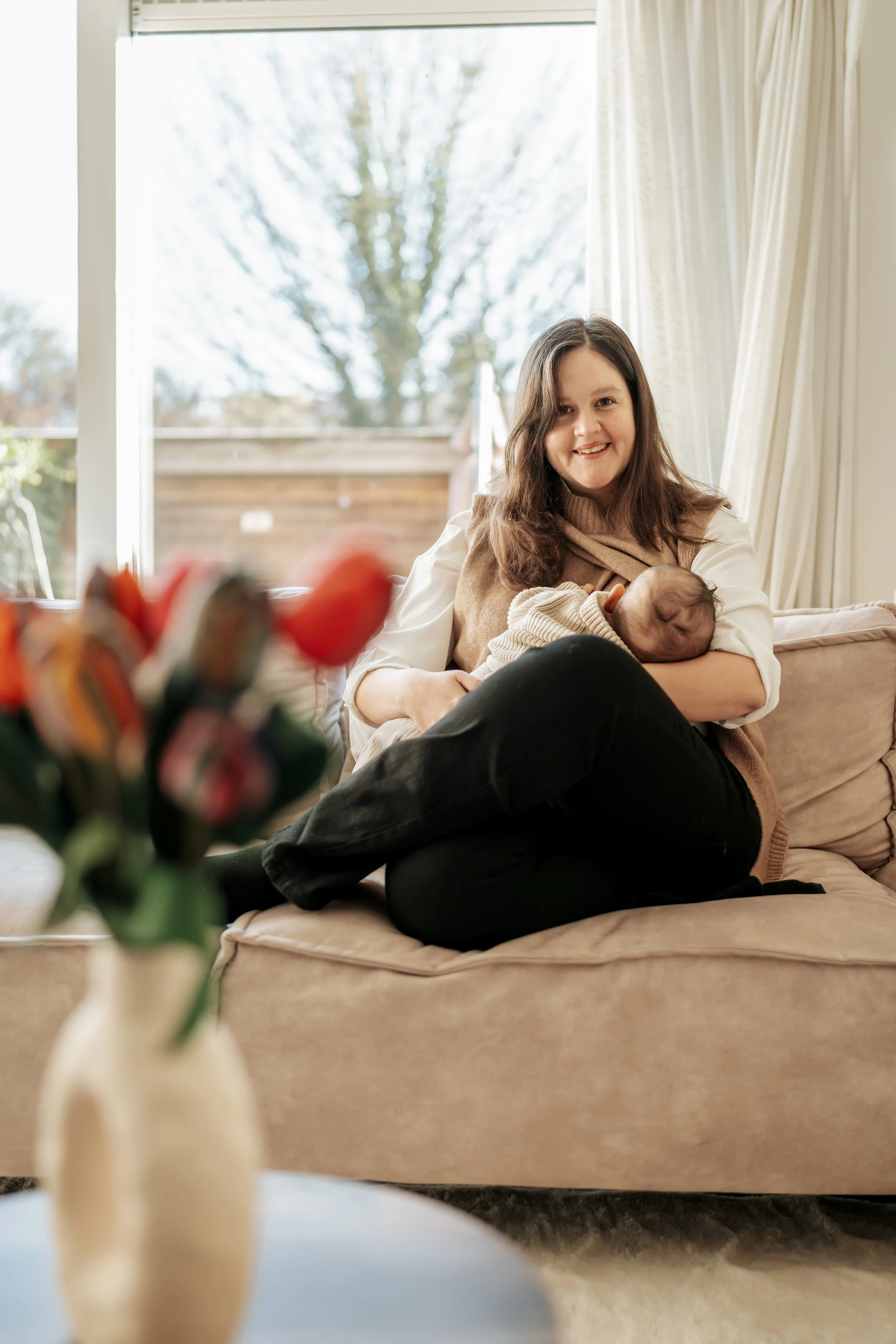 A woman sitting on a beige couch, holding a sleeping baby in her arms, smiling, with a large window and curtains in the background.