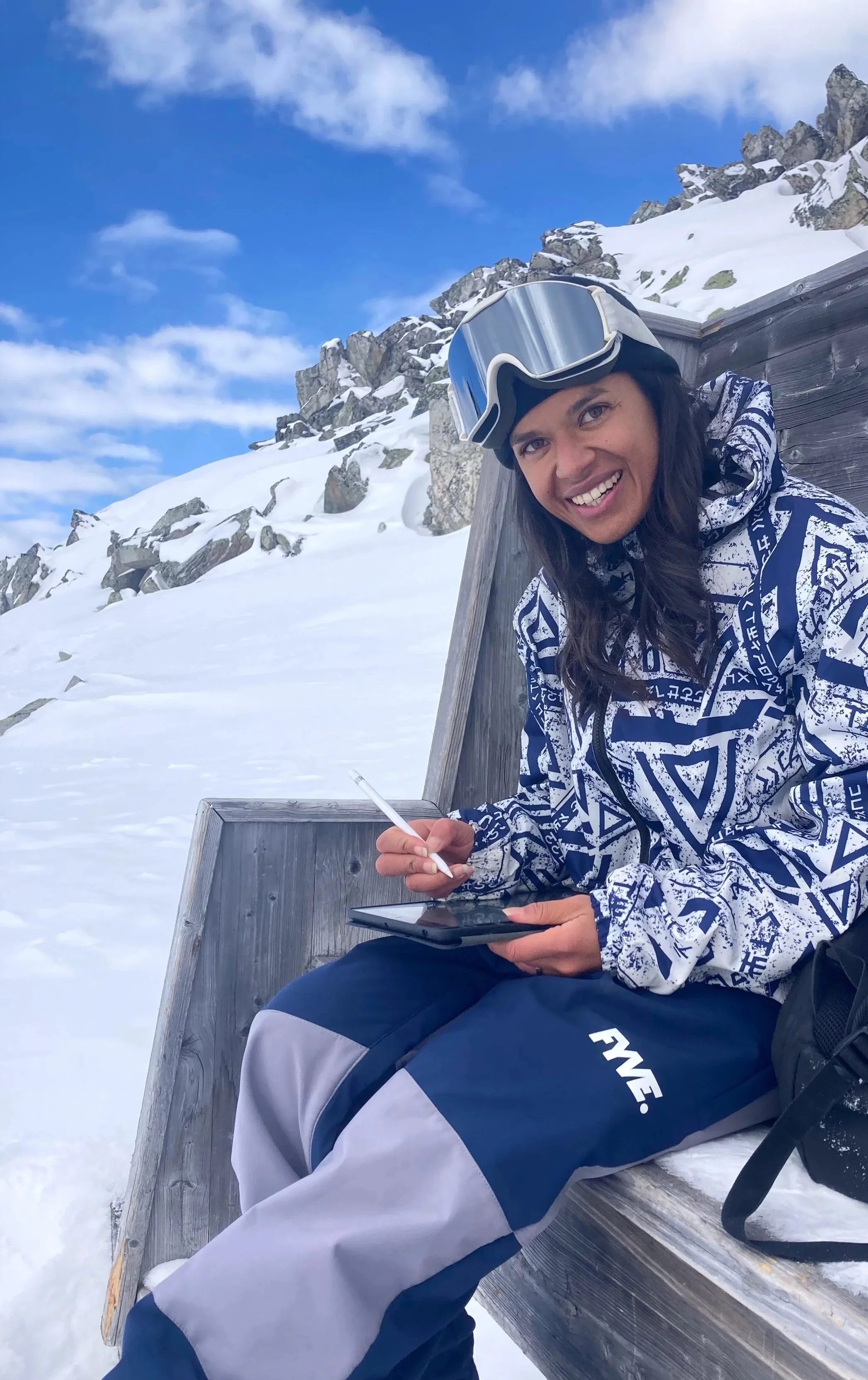 A woman in ski gear sitting on a wooden bench in a snowy mountain landscape, smiling, with ski goggles on her head and a pen in her hand.