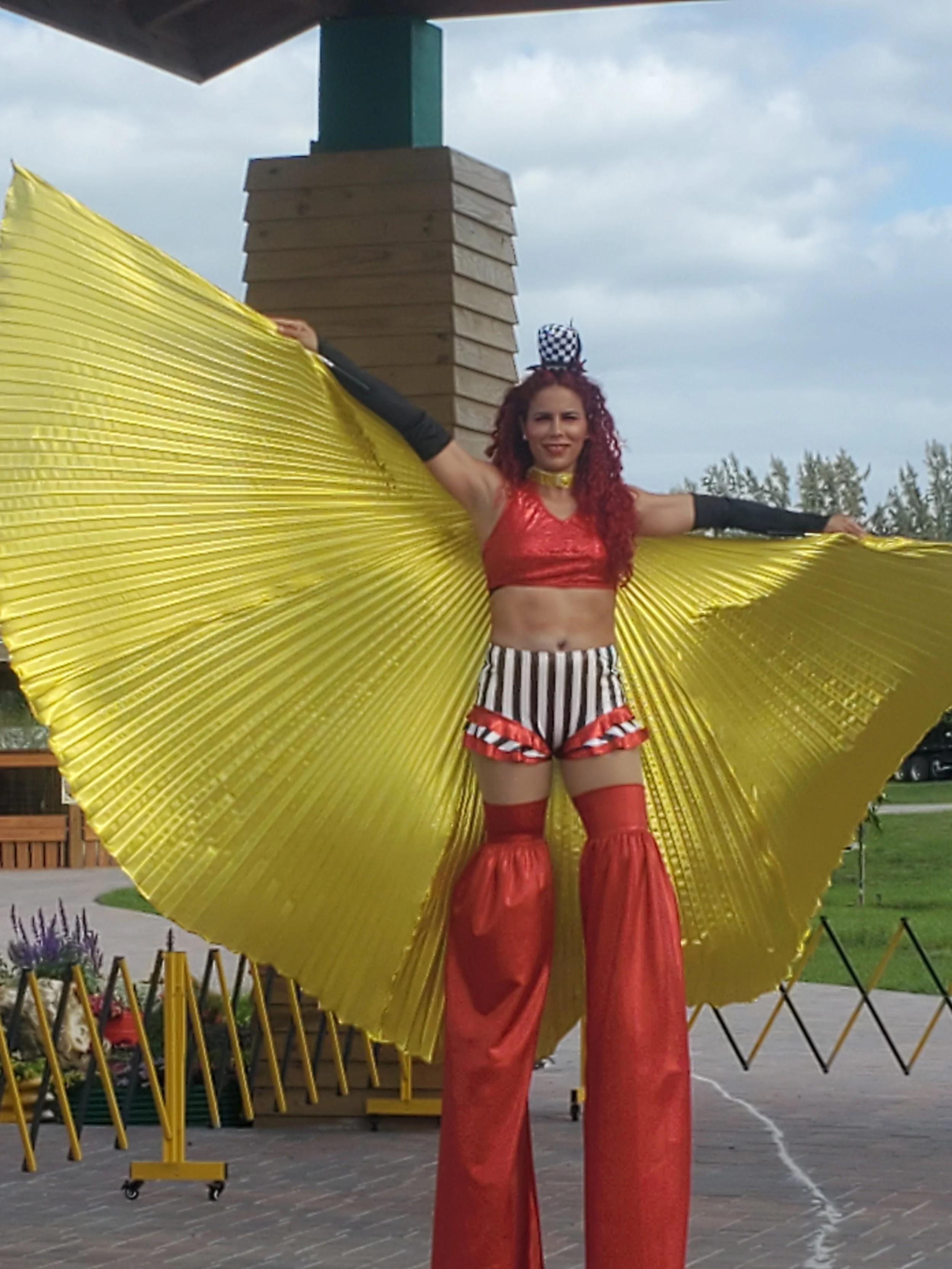 Stilt-walking at the entrance of an event wearing black and white shorts, red sequin cropped top with black gloves , shimmering red stilt-covers, golden butterfly wings, and black and white striped mini top hat headband.