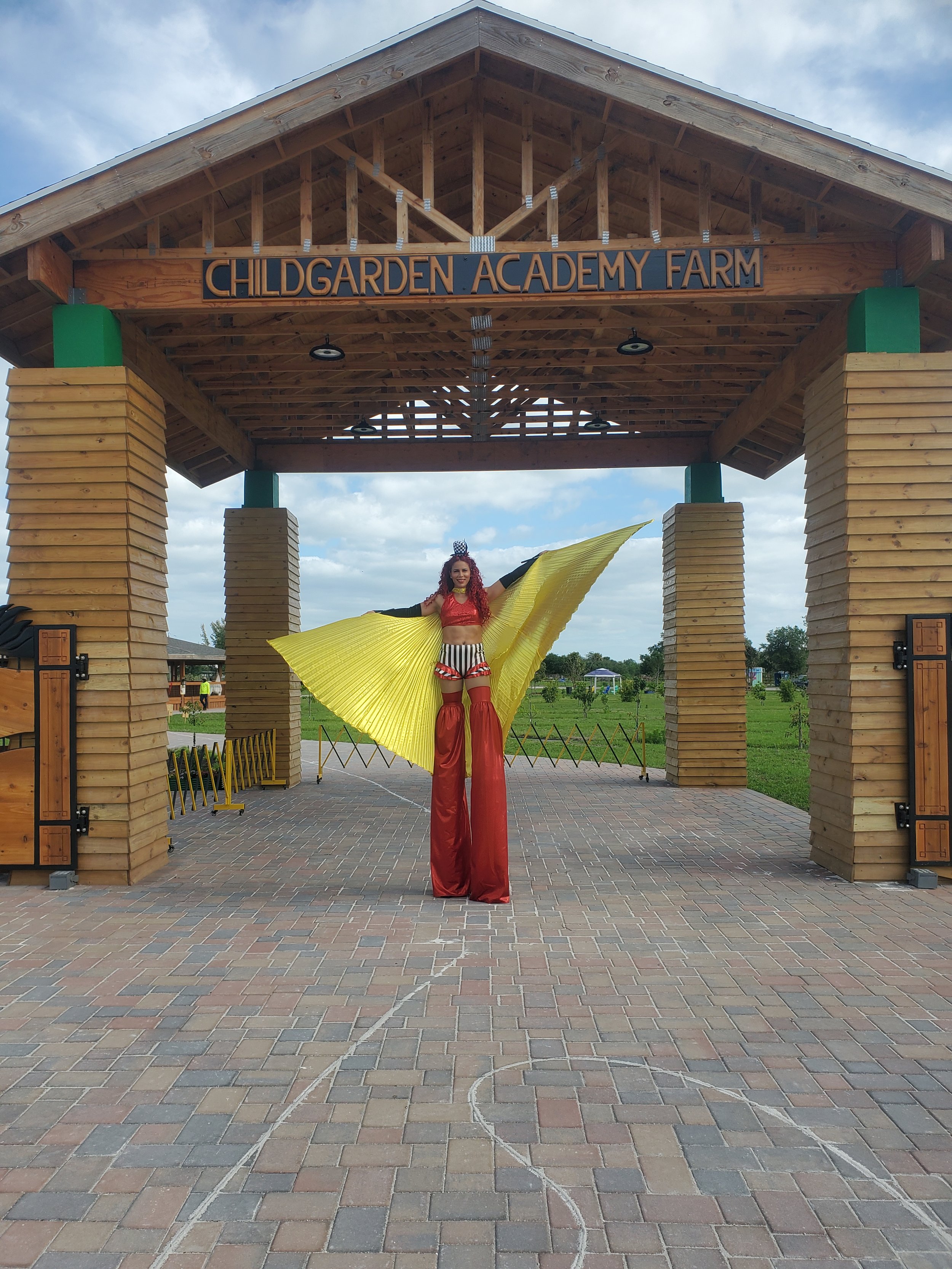 Stilt-walking at the entrance of an event wearing black and white shorts, red sequin cropped top with black gloves , shimmering red stilt-covers, golden butterfly wings, and black and white striped mini top hat headband.