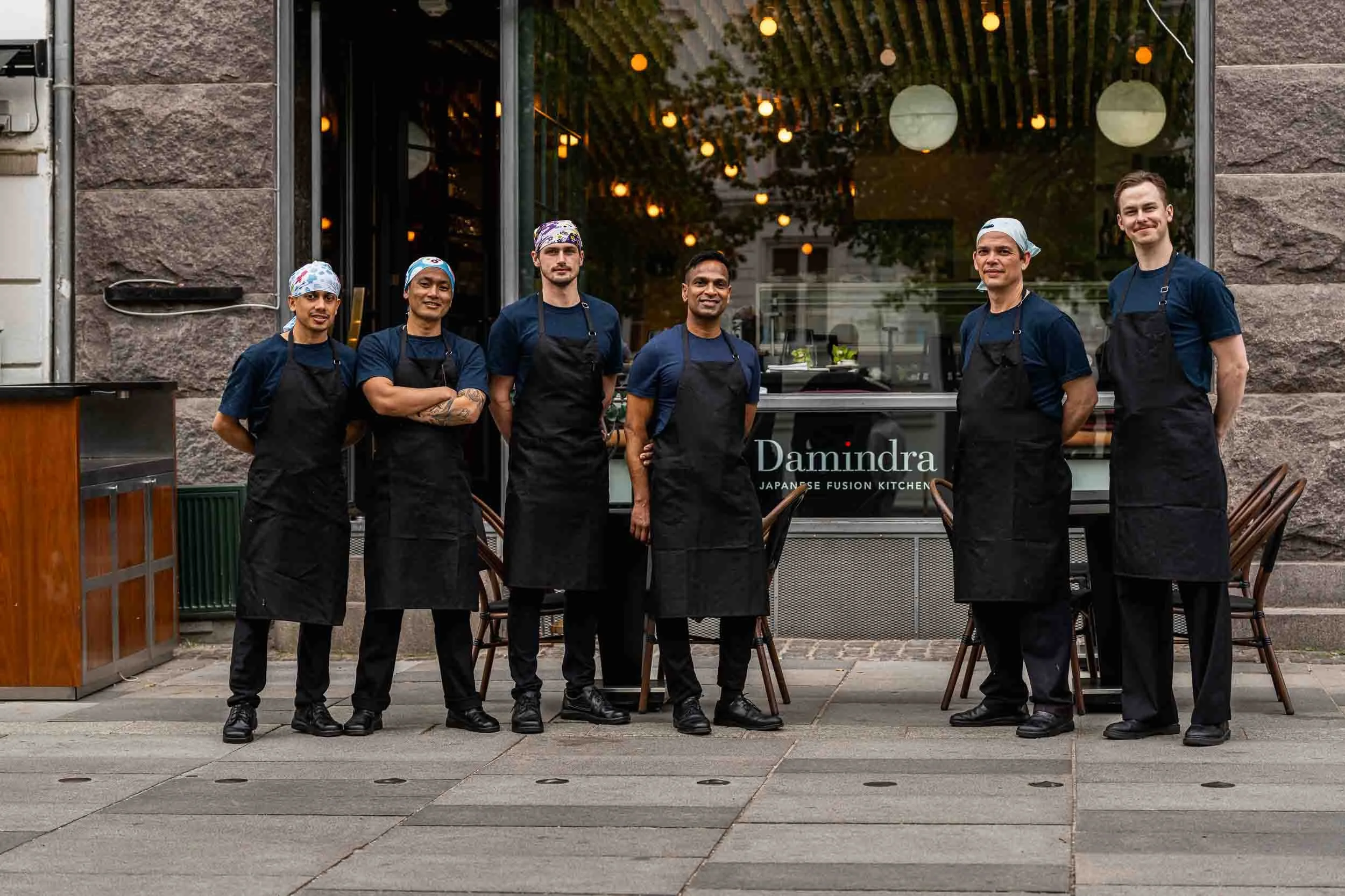 Seks kokke står udenfor en japansk fusion-restaurant, alle iført blå t-shirts, sorte forklæder og hovedbeklædning, foran et vindue med restaurantenavnet Damindra.