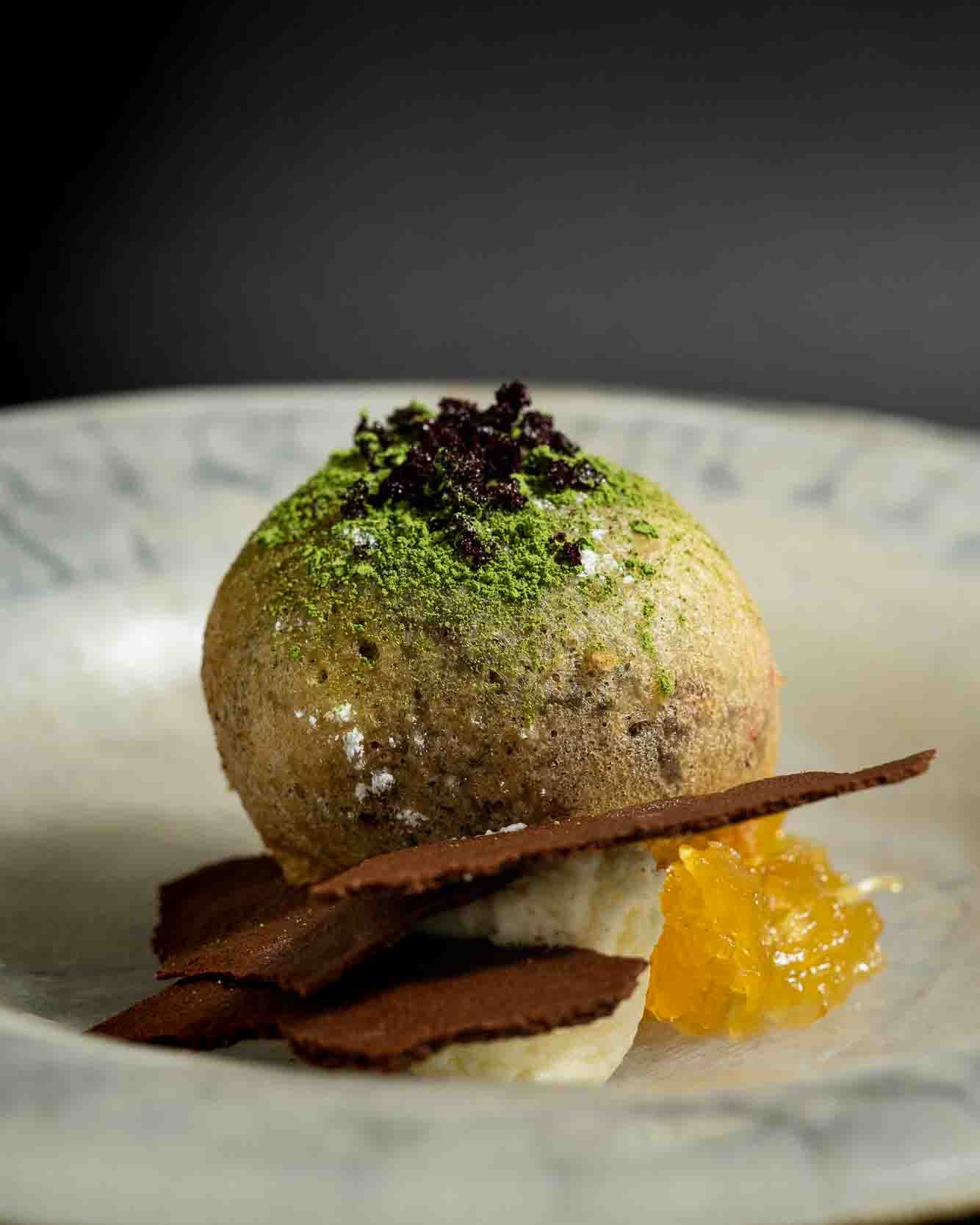 En moderne dessert med en stor, rund kugle dækket af grøn pulver, pyntet med små sorte korn og serveret på en tallerken med lag af chokolade, skefuld marmelade og mousse.
