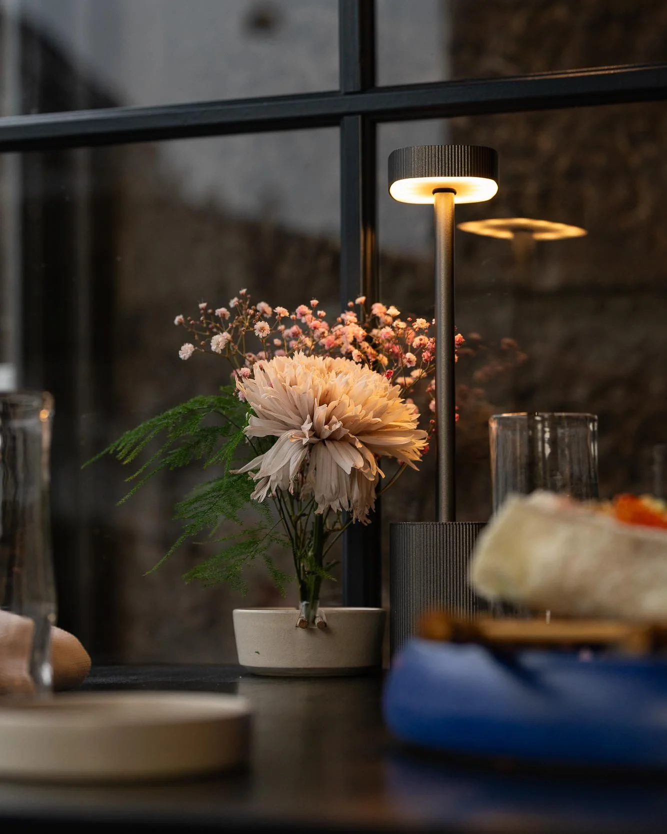 En bord med en vase med lyserøde blomster, en bordlampe, en tallerken og en glas vinstemperatur, sat op ved en vindueskarm.
