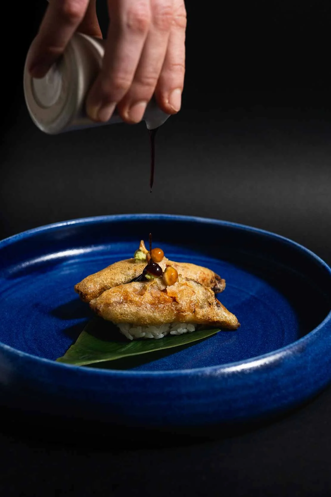 En person hælder en mørk sauce over en japansk sushi med sprød fisk på en blå tallerken med grøn blad under.
