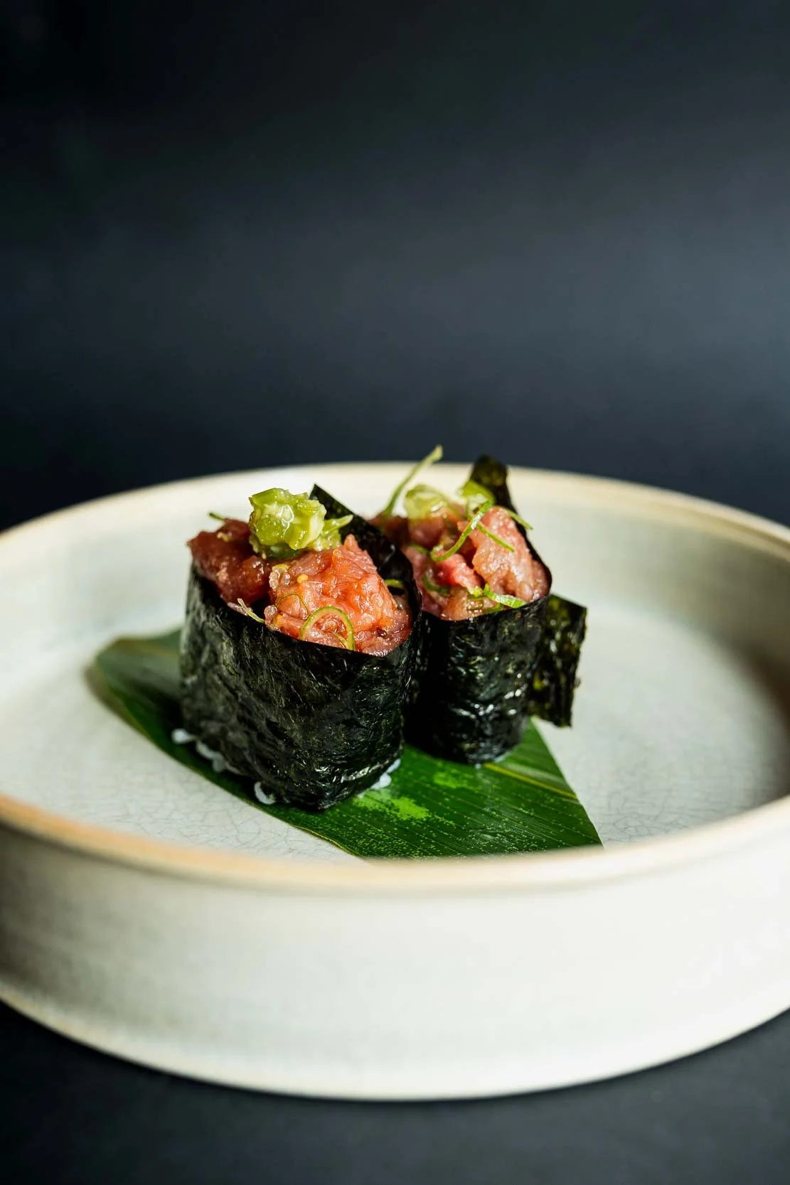 To sushi ruller med rå fisk, indpakket i tang, lagt på en grønne blad på en hvid tallerken.