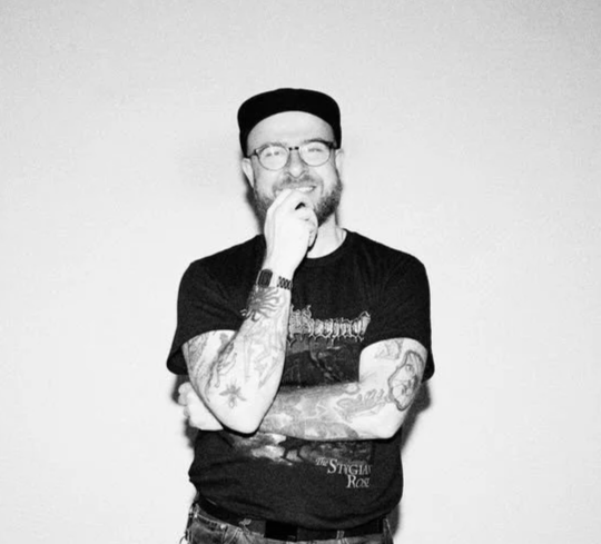 A man with tattoos on his arms, wearing glasses, a black t-shirt, and a black beanie, smiling and standing with his hand near his chin against a plain background.