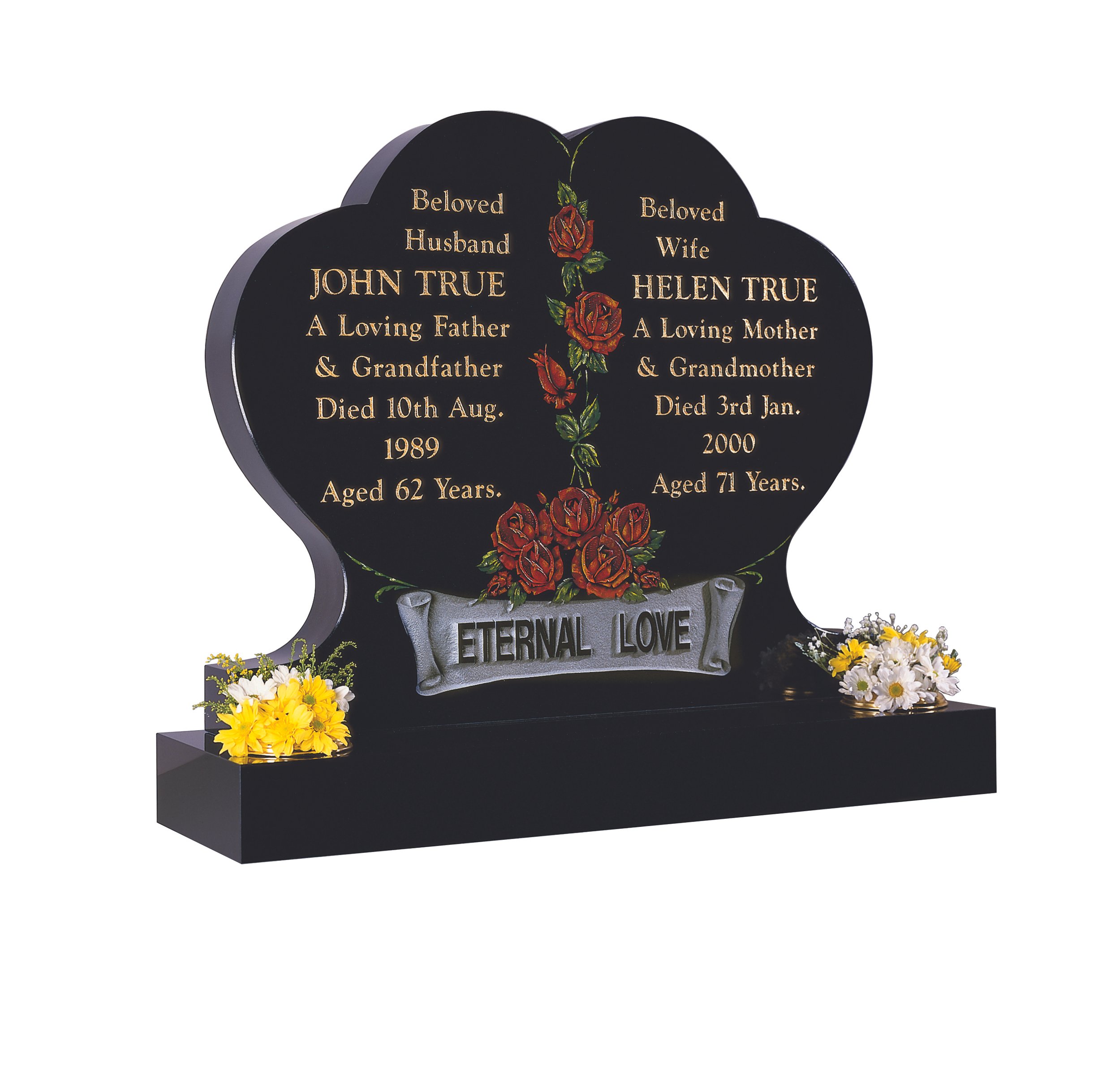 A black double heart-shaped tombstone with red roses and green leaves engraved in the center. The tombstone contains the names and details of John True and Helen True, along with their death dates and ages. The phrase 'ETERNAL LOVE' is engraved on a ribbon at the bottom, and there are yellow and white flower arrangements on each side of the tombstone.