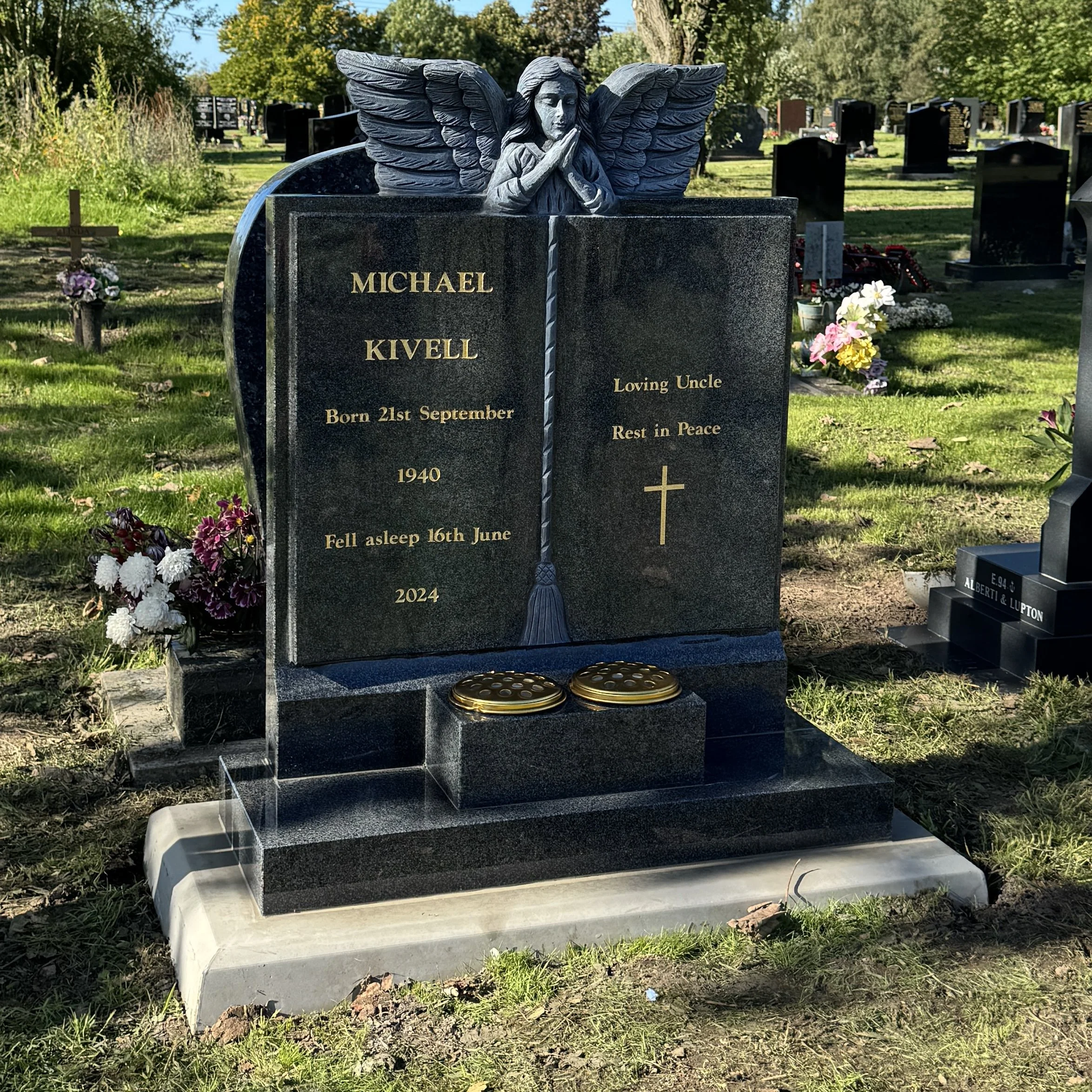 Black granite tombstone with an angel sculpture on top, depicting a praying angel with wings, in a cemetery. The tombstone commemorates Michael Kivell, born September 21, 1940, and passed away June 16, 2024. It includes inscriptions 'Loving Uncle' and 'Rest in Peace' with a cross symbol. There are two round gold-colored ornaments at the base of the tombstone.