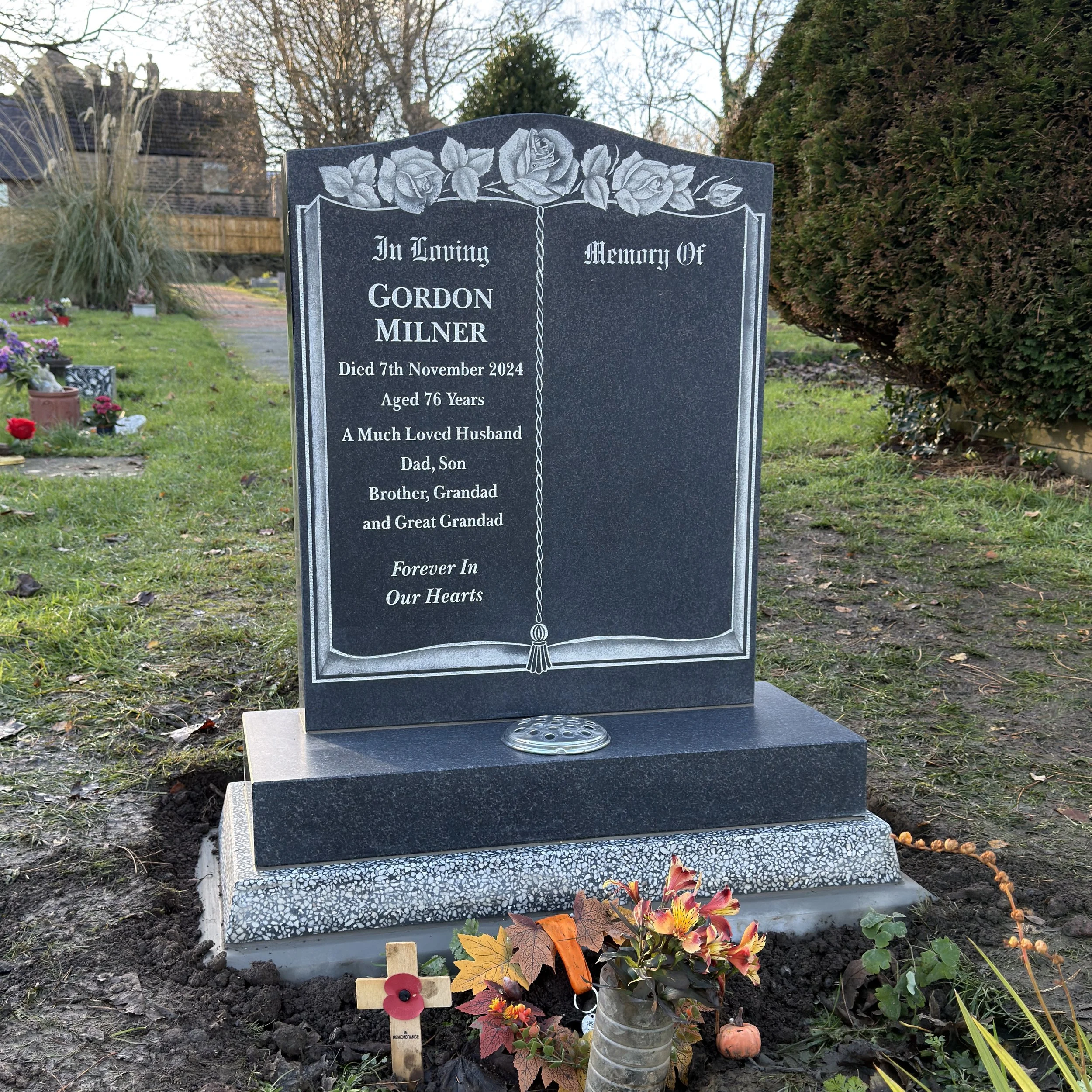 A black granite headstone with engraved roses at the top. The inscription commemorates Gordon Milner, who died on November 7, 2024, at age 76. The stone features a decorative border and a chain design in the center. There are flowers, a small wooden cross, and a miniature pumpkin at the base of the headstone. The background shows a grassy area with other graves and trees.