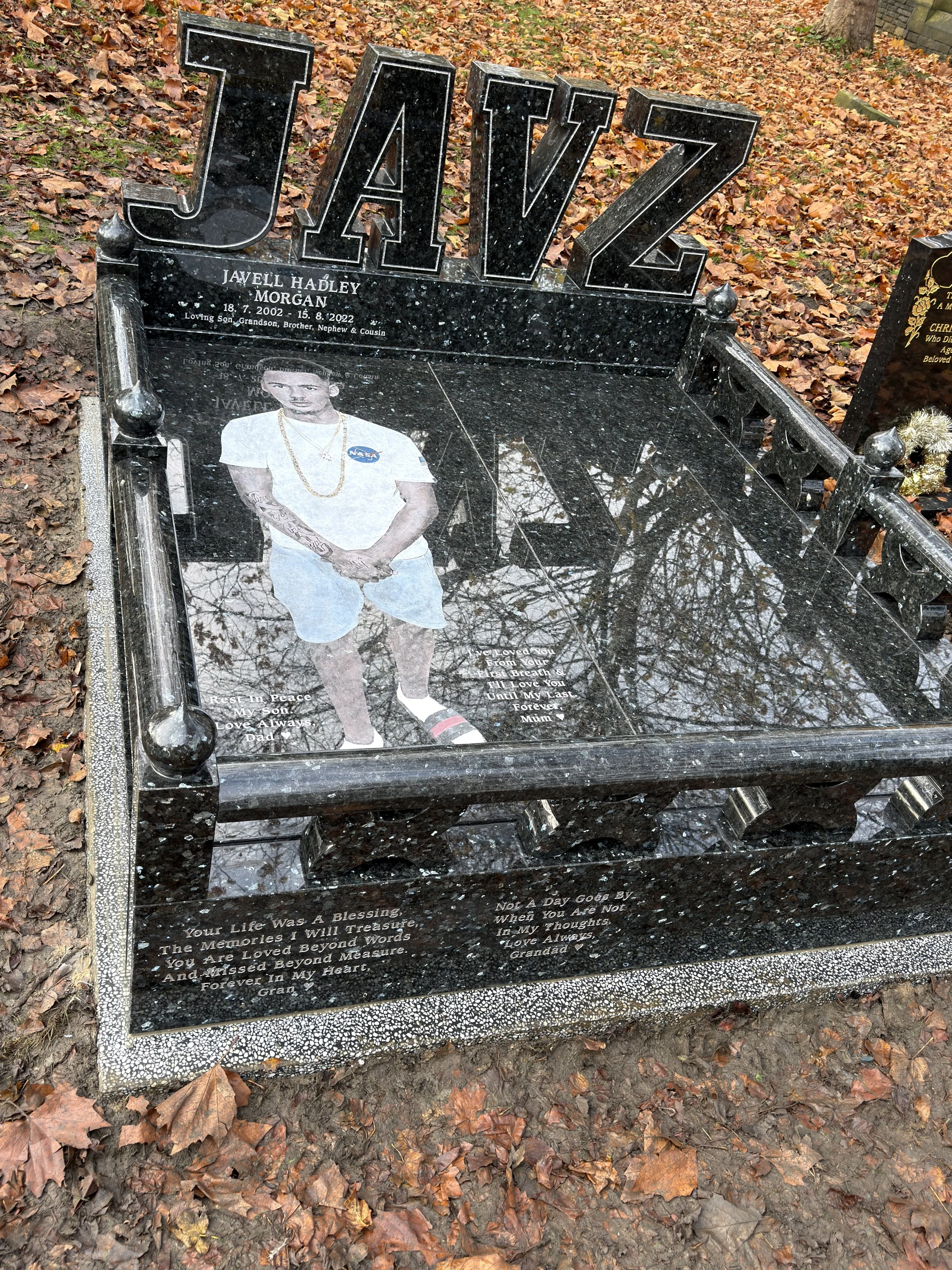 A gravestone with the name 'JAVZ' at the top and a photo of a young man in casual clothing. The gravestone is black with engraved writings, surrounded by fallen autumn leaves.