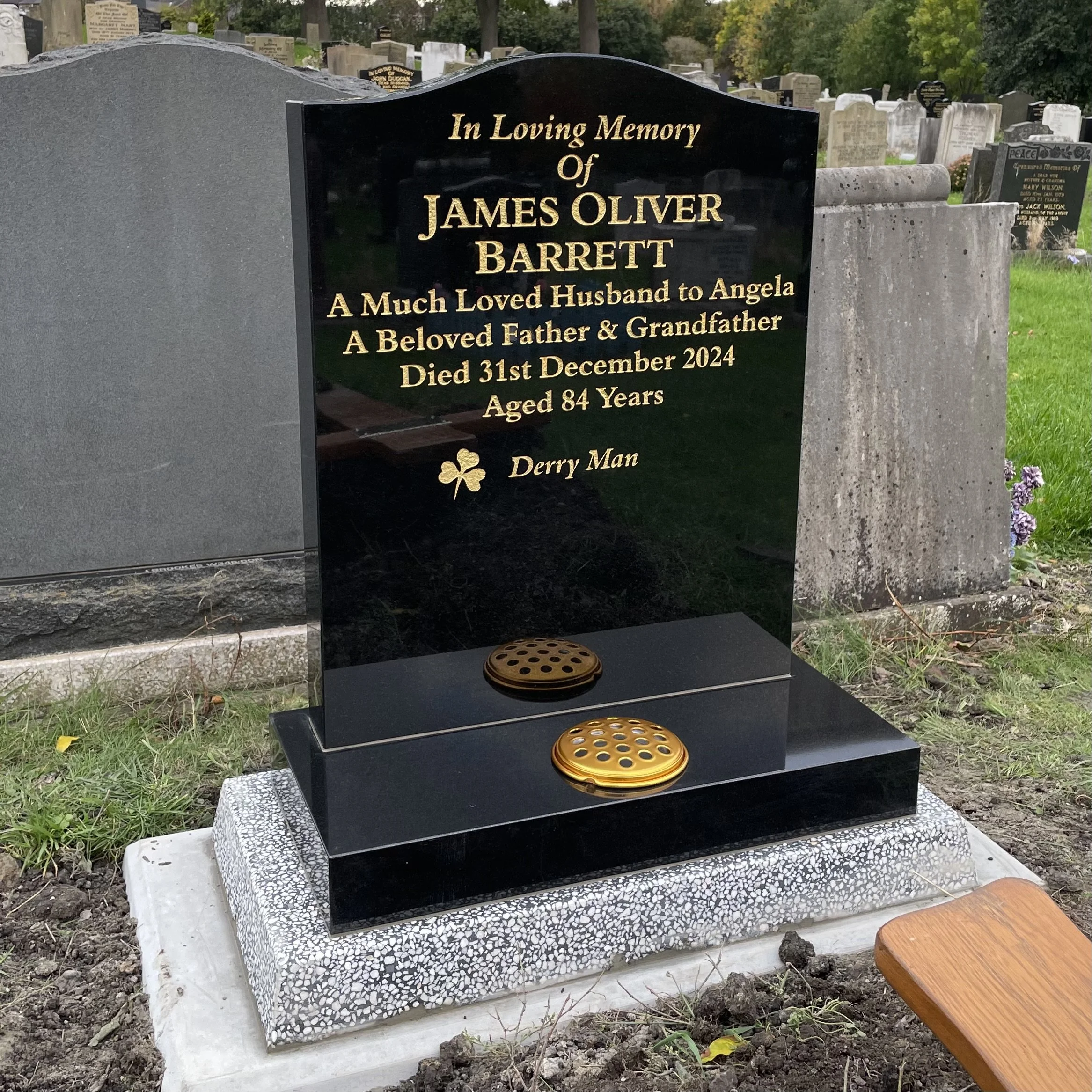 Black granite headstone with gold inscriptions in a cemetery, honoring James Oliver Barrett, a beloved husband, father, and grandfather who died at age 84 on December 31, 2024. The headstone features a four-leaf clover symbol and two commemorative discs at the base.
