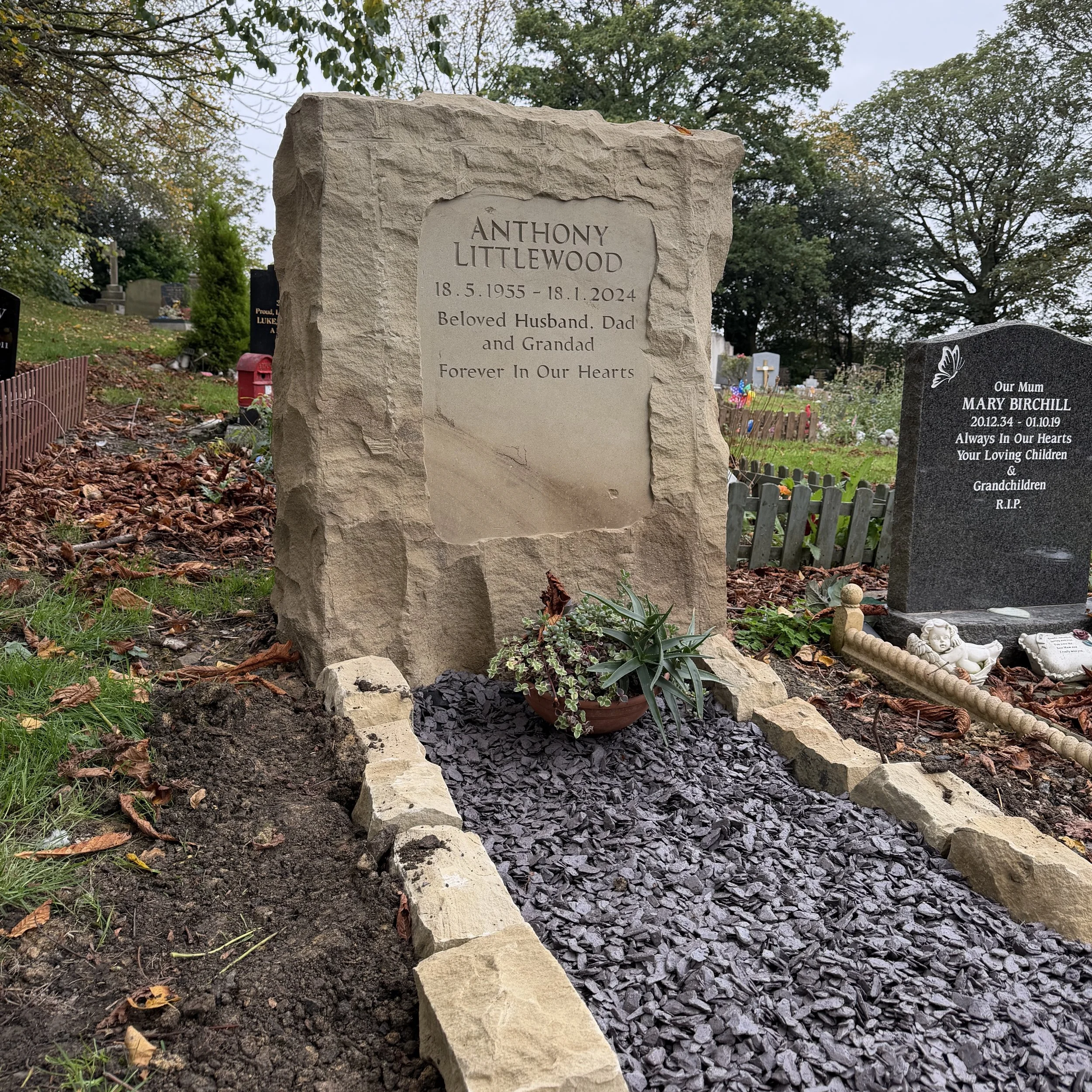 Gravestone in a cemetery for Anthony Littlewood, born May 18, 1955, died January 18, 2024, with the words 'Beloved Husband, Dad, and Grandad, Forever In Our Hearts', surrounded by fallen leaves and a small garden with a potted plant.