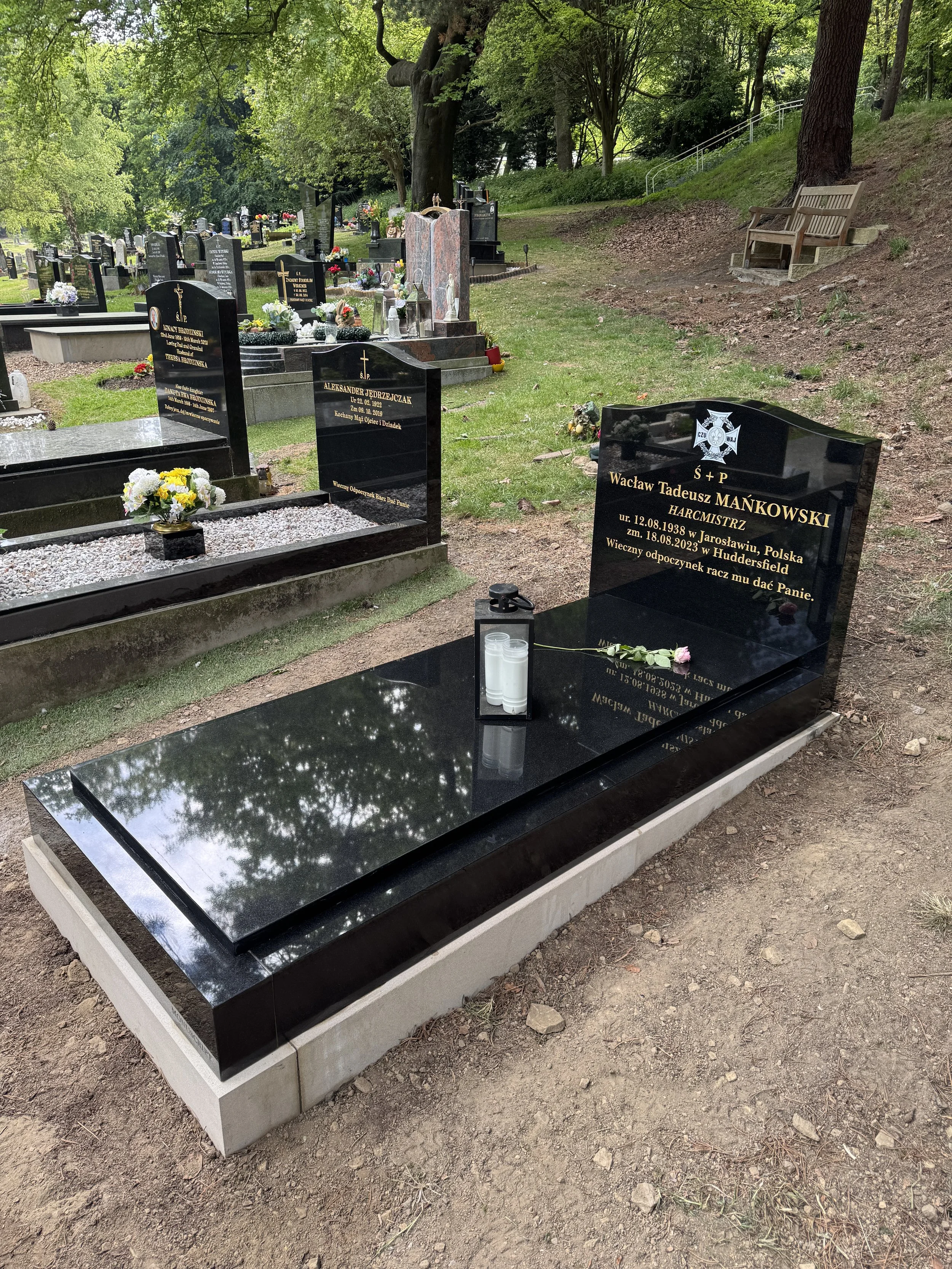 A black granite gravestone with yellow writing, decorated with a pink rose and a lantern containing white candles, located in a cemetery with other gravestones and trees in the background.