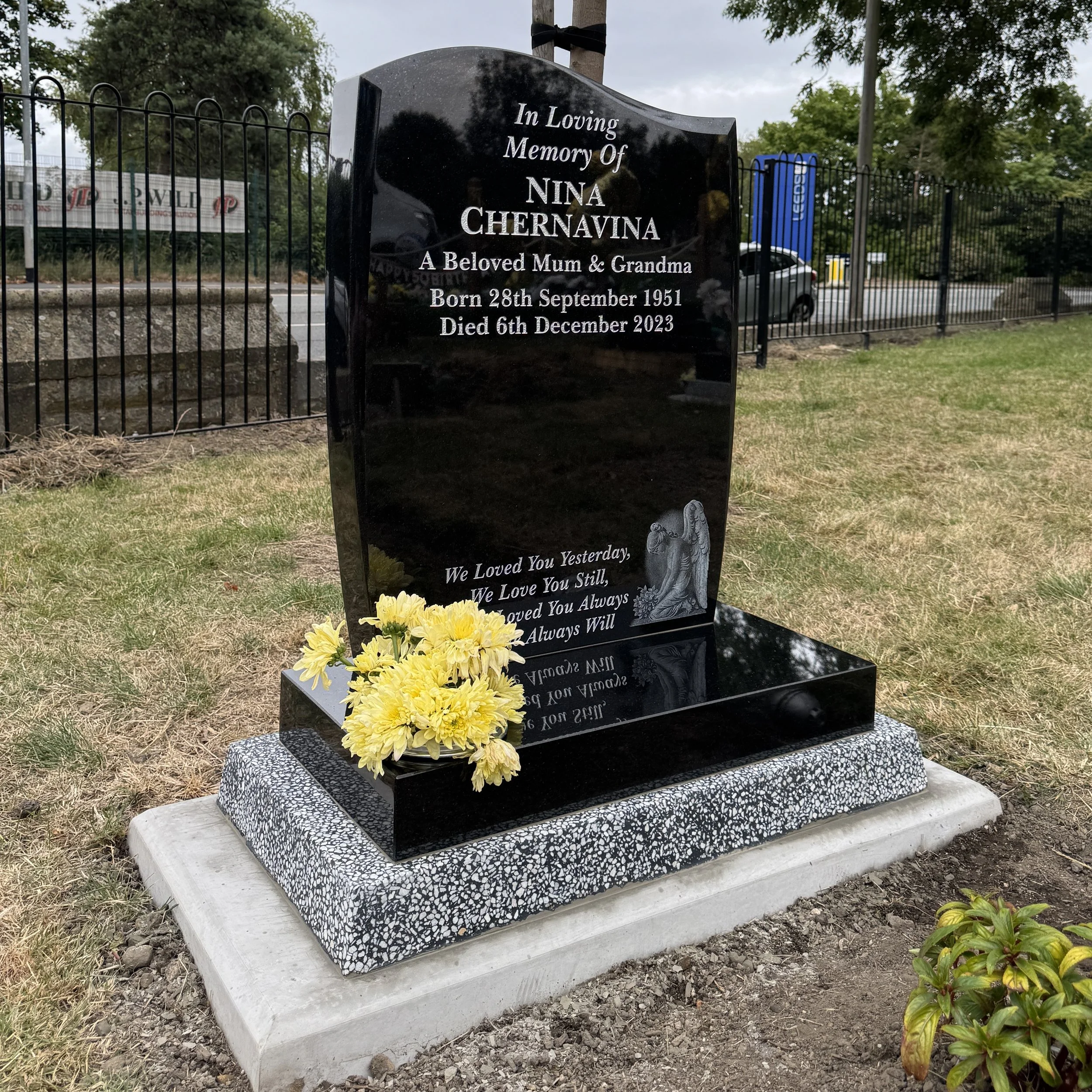 A black polished granite headstone for Nina Chernavina, with yellow flowers on the base, in a cemetery with grass and trees, surrounded by a black metal fence.