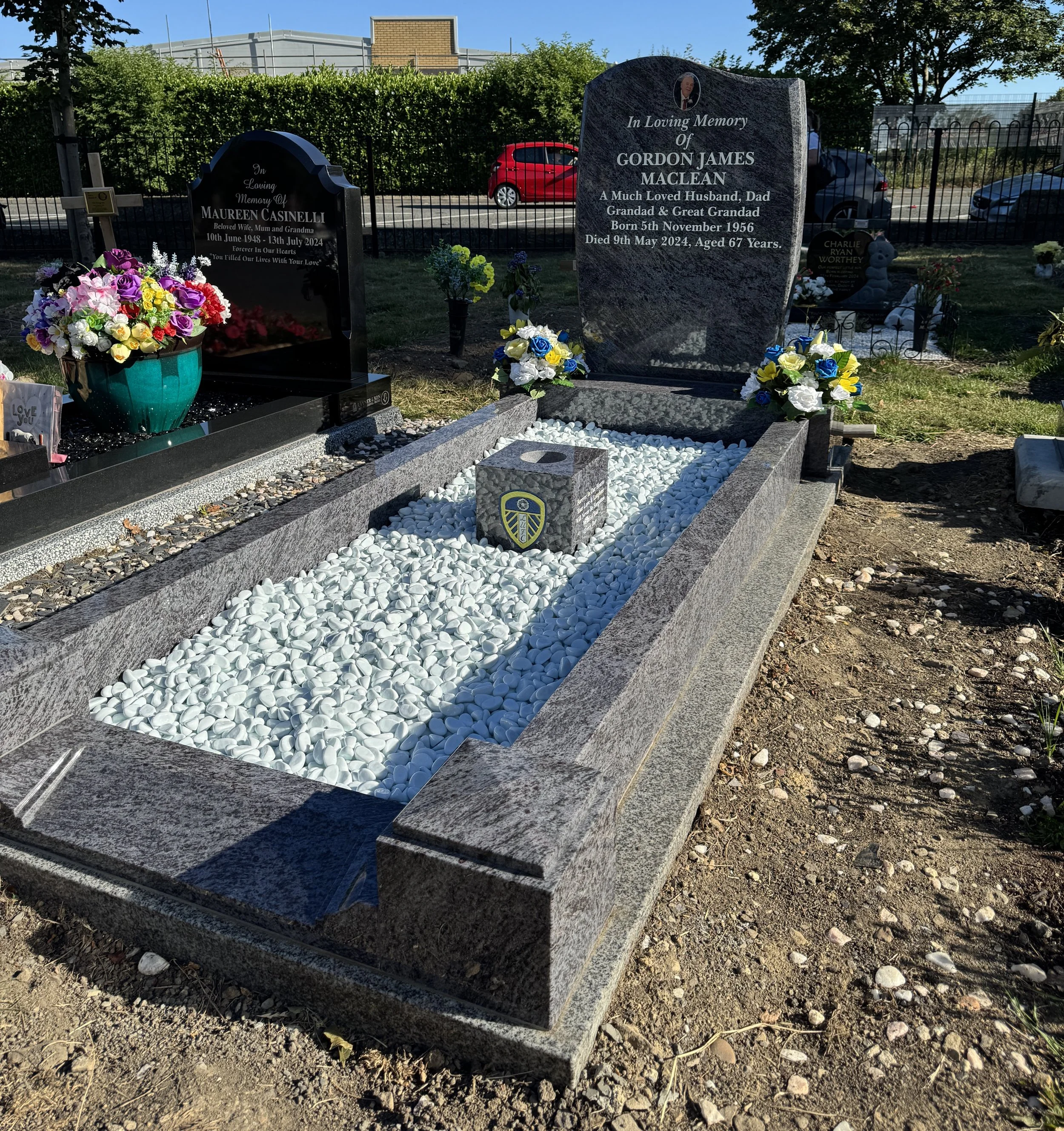 Grave with a granite headstone for Gordon James Maclean, surrounded by white pebbles and colorful floral arrangements, in a cemetery with greenery and other graves in the background.