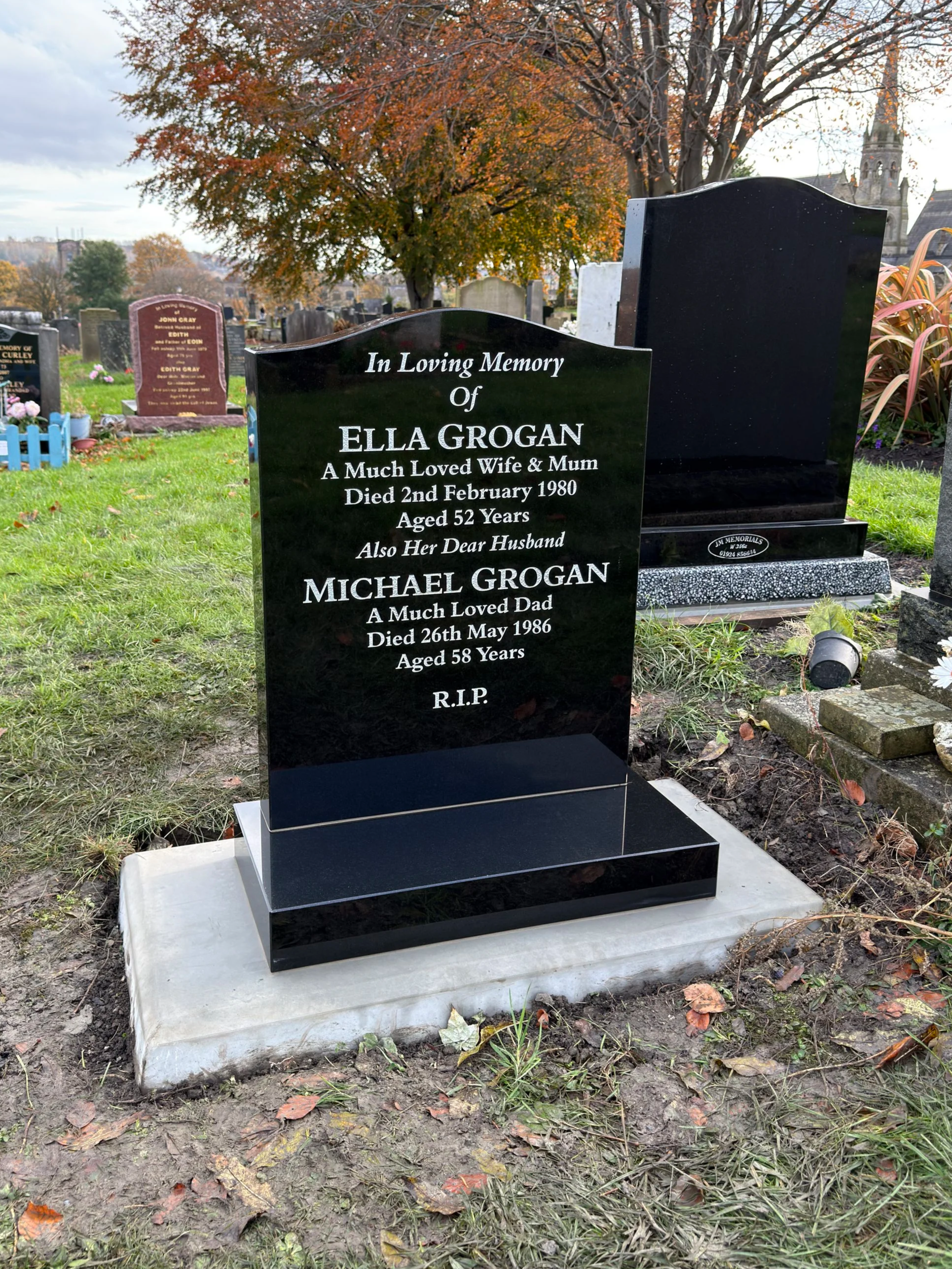 A black polished headstone in a cemetery with an inscription in memory of Ella Grogan and Michael Grogan, with trees and other grave markers in the background.
