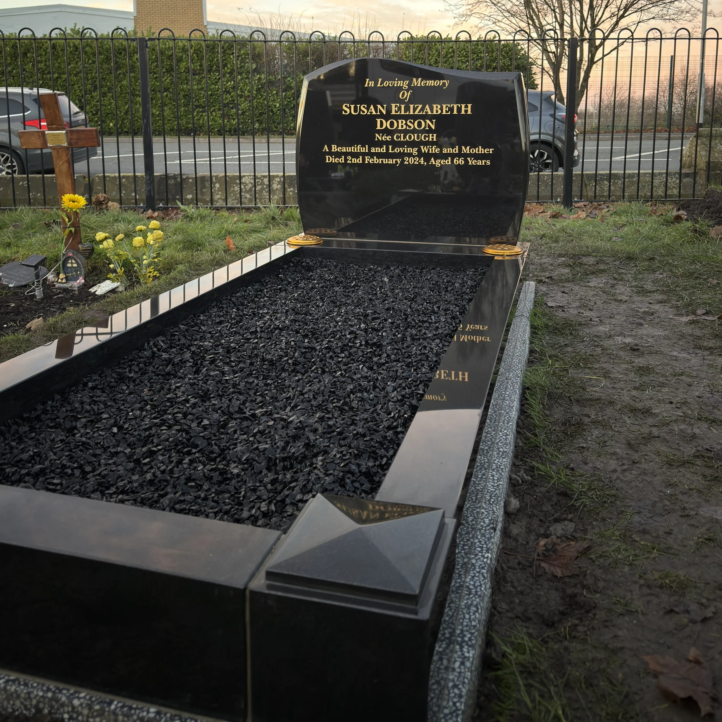 Grave site with black polished headstone reading 'In Loving Memory of SUSAN ELIZABETH DOBSON née CLOUR, A Beautiful and Loving Wife and Mother, Died 2nd February 2024, Aged 66 Years.' There is a small yellow sunflower and other floral decorations next to the grave, a wooden cross, and a little decorative item beneath the cross. A black iron fence surrounds the cemetery, with parked cars and trees visible in the background during either dawn or dusk.